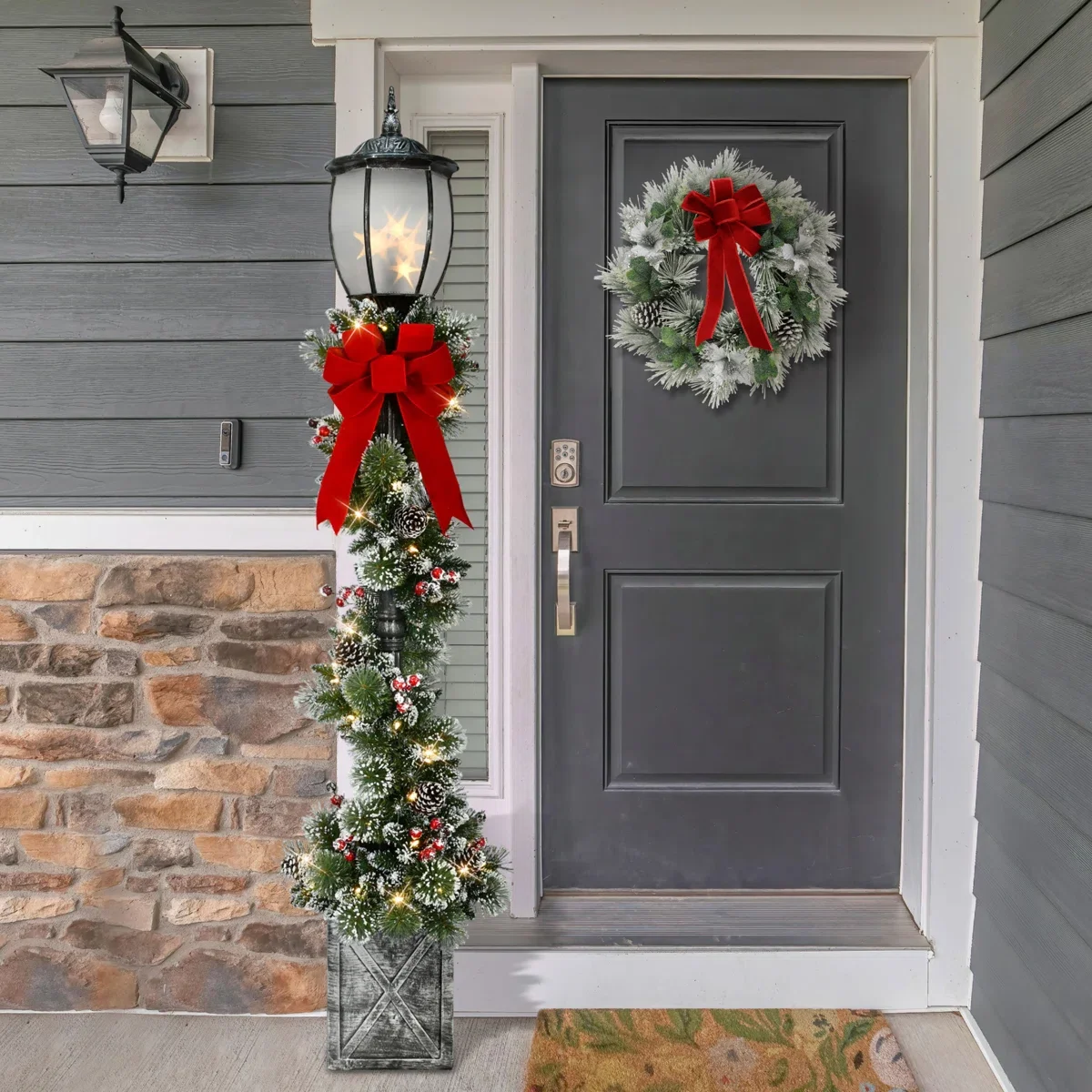 Front door decorated for Christmas with a wreath on the door, a lit lamp post with a red bow and ornaments, and a small Christmas tree in a grey wooden planter.