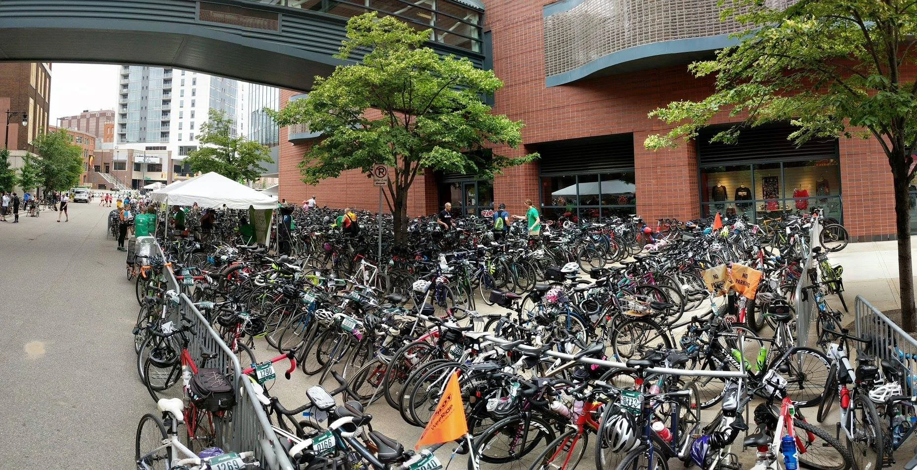 A large outdoor bike parking area filled with bicycles, with people walking and biking nearby, tall buildings in the background, and trees providing shade.