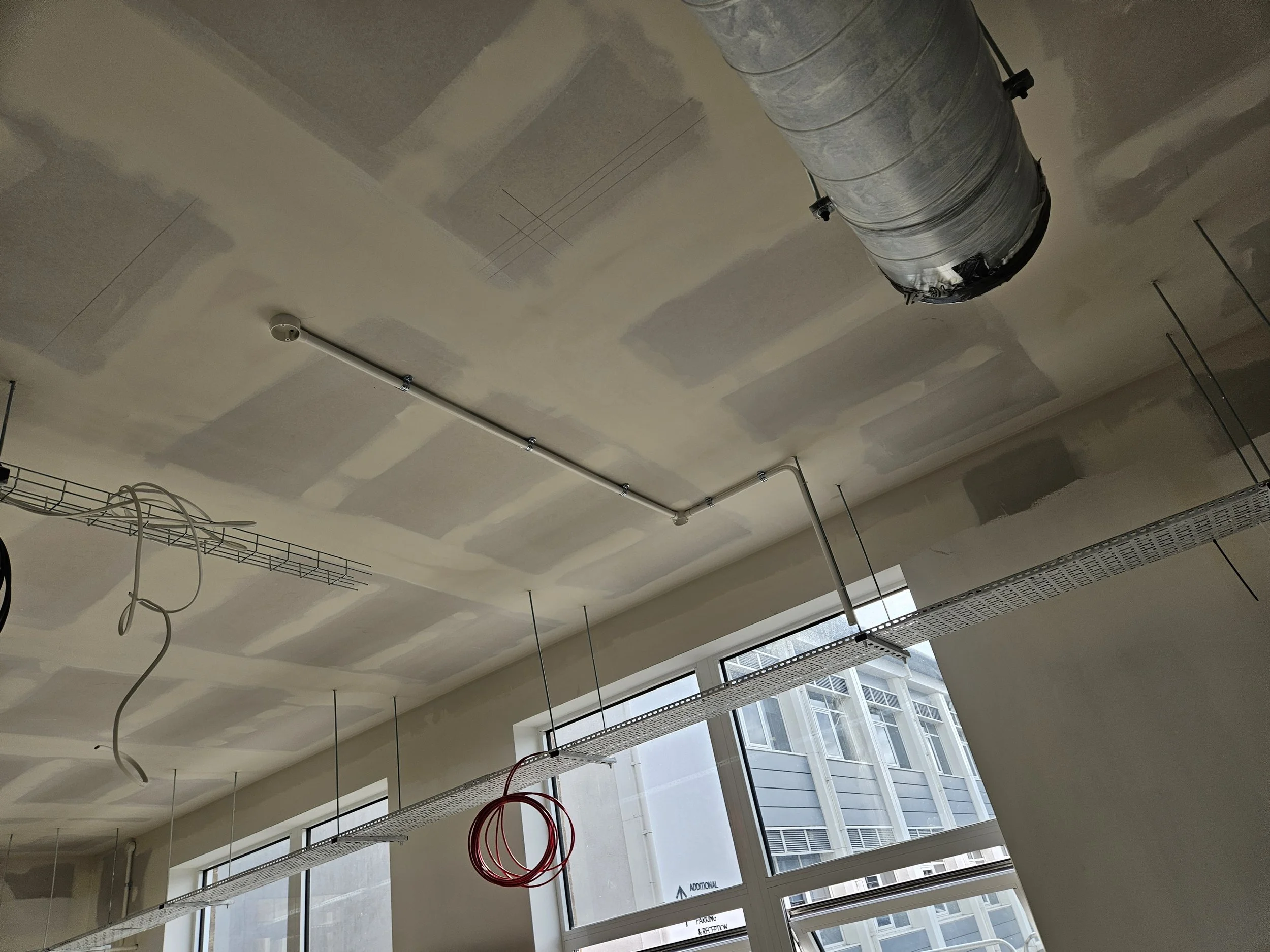 Interior of a building under construction with exposed ductwork, electrical wiring, and a suspended cable tray, with windows showing neighboring buildings.