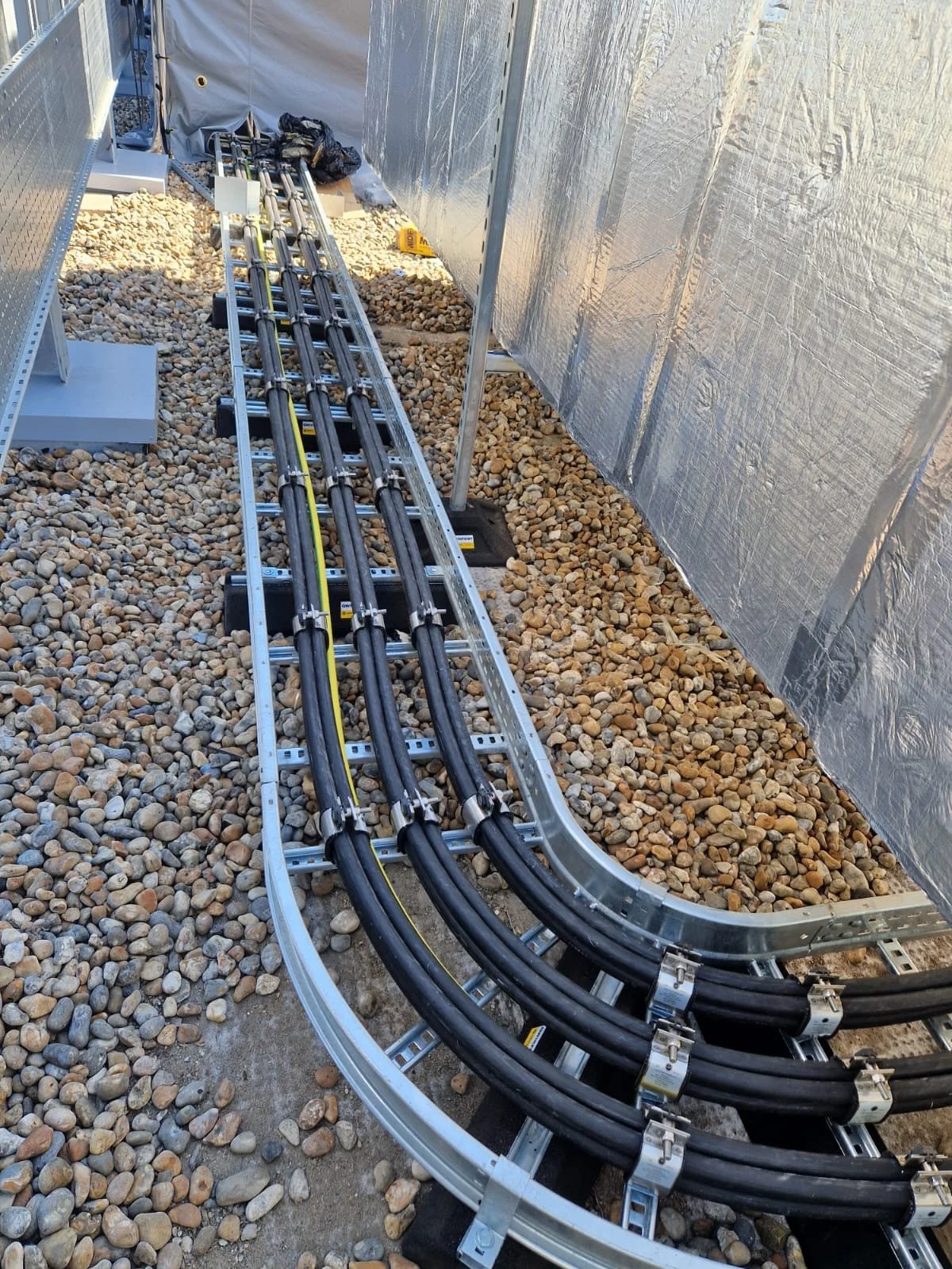 Close-up of a metal cable management tray with five thick black cables secured by metal clamps, situated on a gravel ground next to a reflective wall, with additional construction tools and materials in the background.
