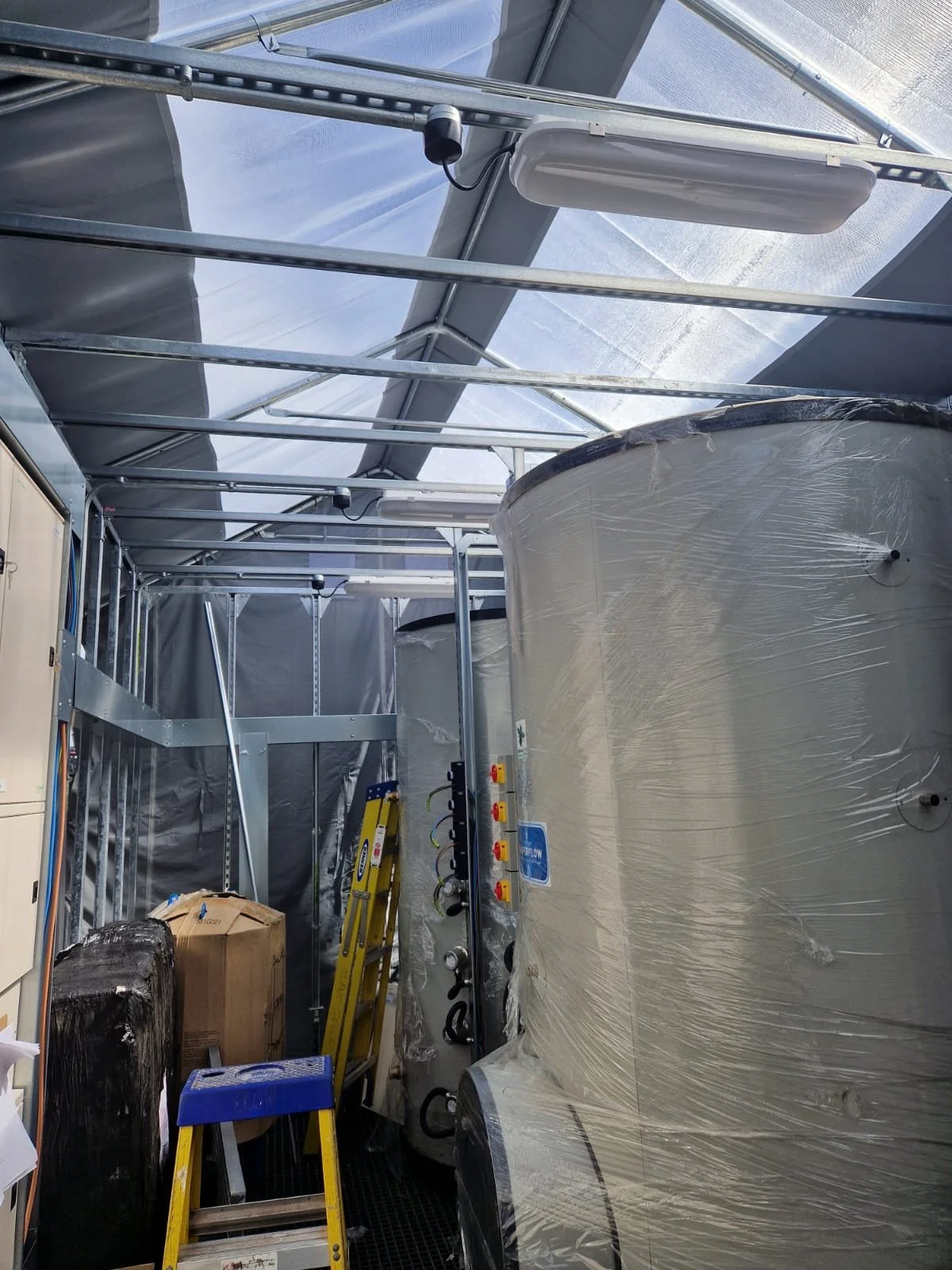 Inside a greenhouse with large wrapped water tanks, a yellow step ladder, and a blue stool.