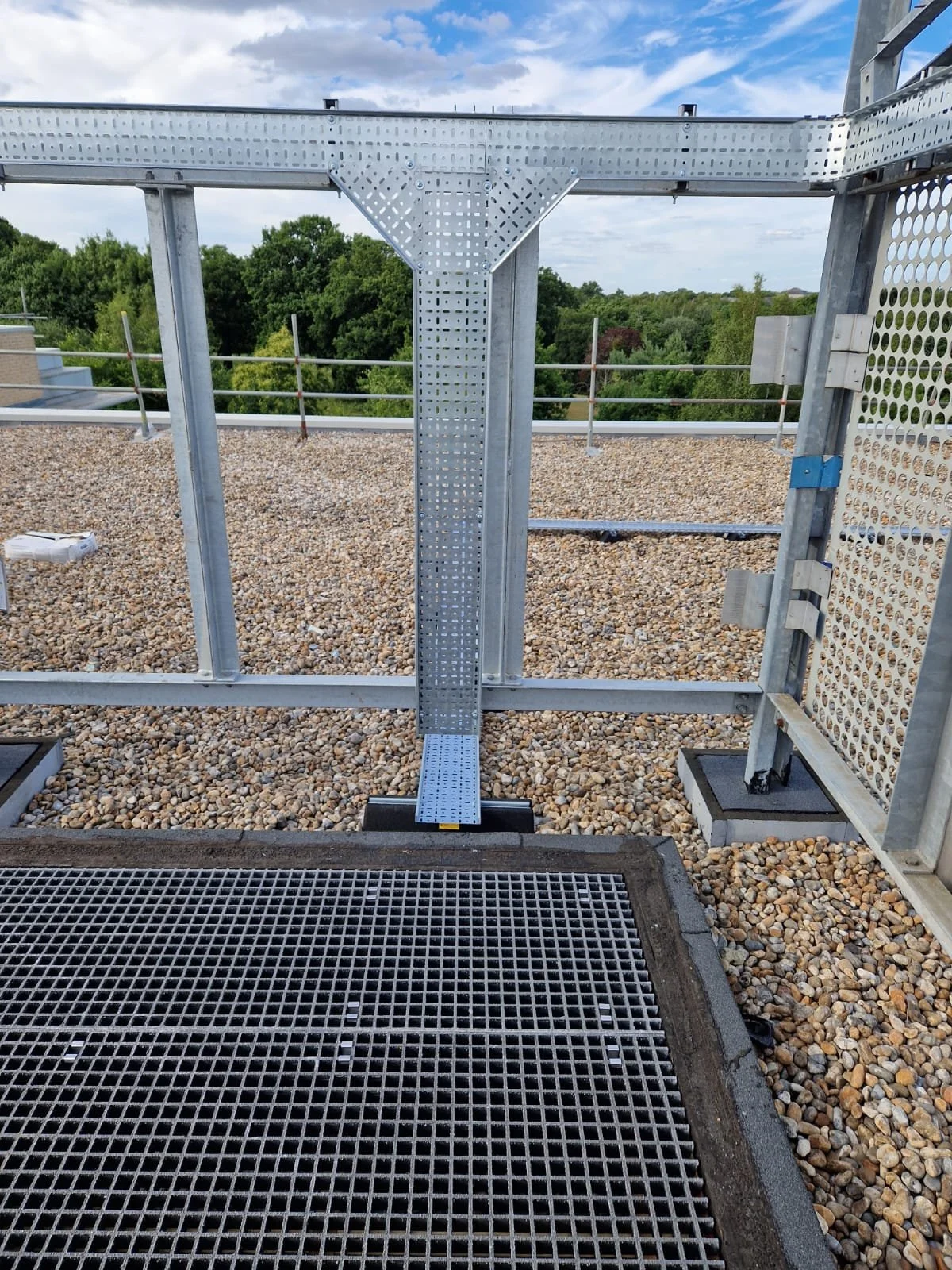 Metal structure with a perforated vertical beam, a grated floor, and a protective mesh wall, located outdoors on a rooftop with trees and a cloudy sky in the background.