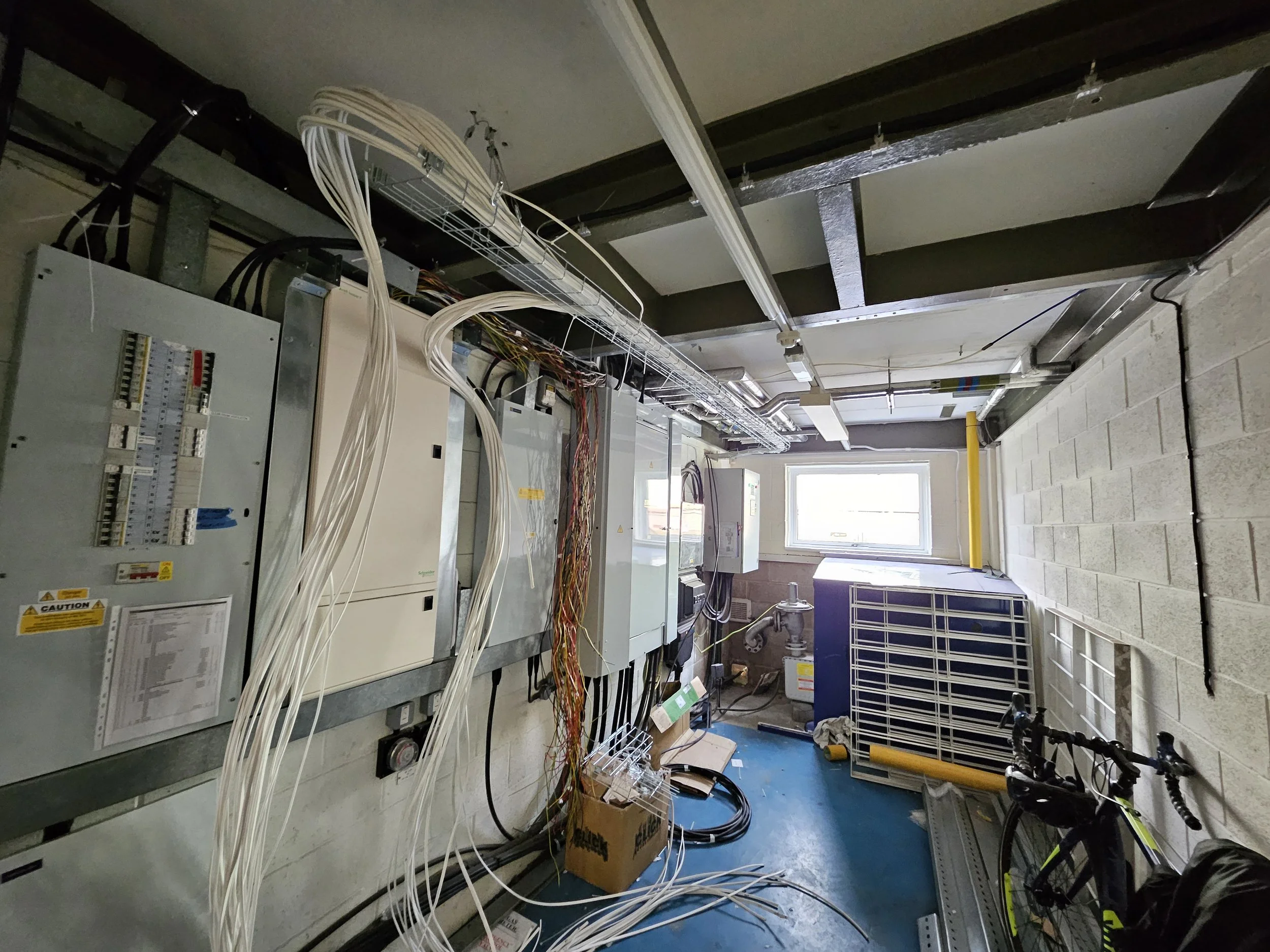 Electrical wiring and panels in a utility room with a window, a bike, and construction supplies.