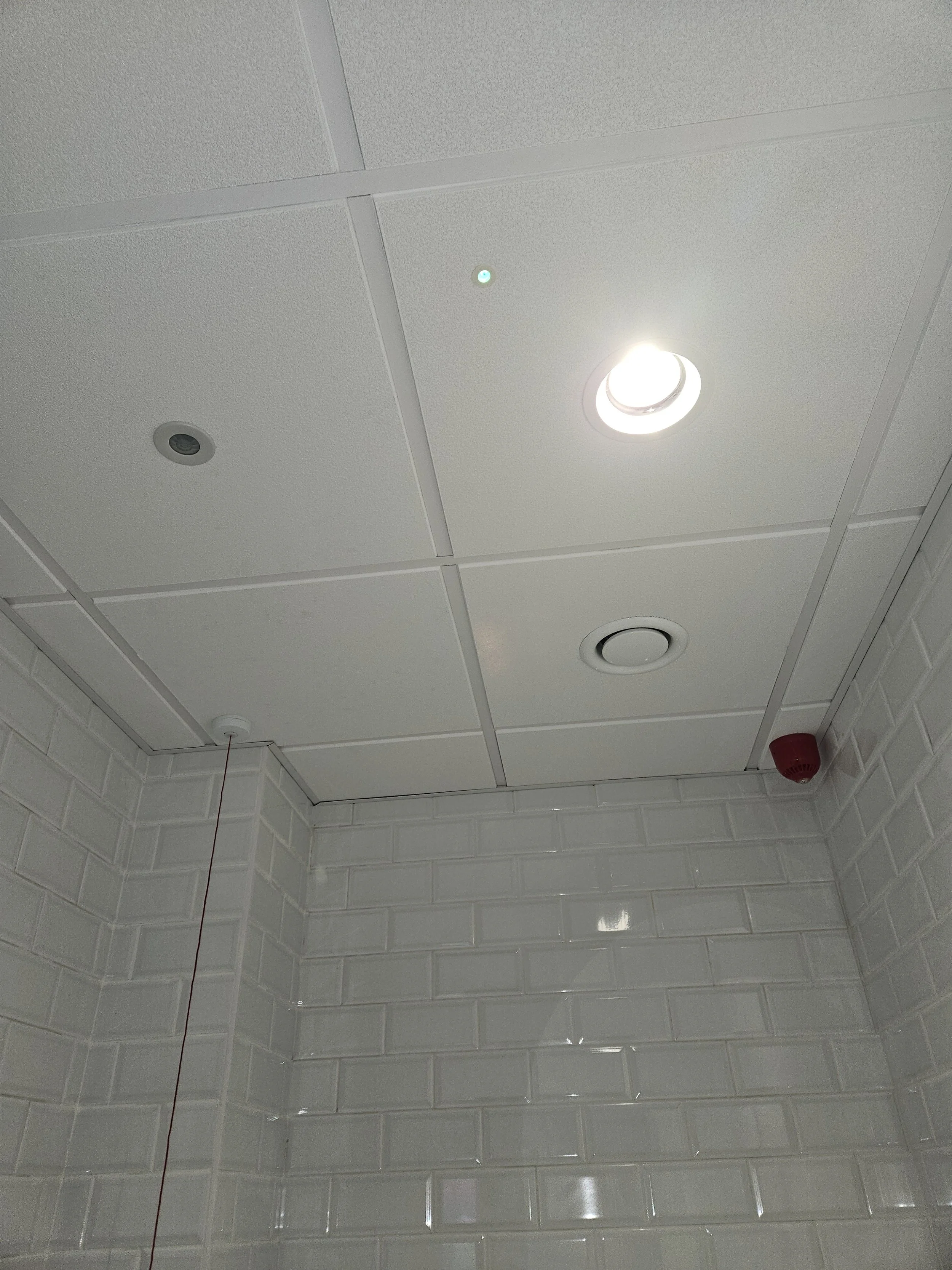 Ceiling with recessed lighting, smoke detector, and red alarm on tiled wall.
