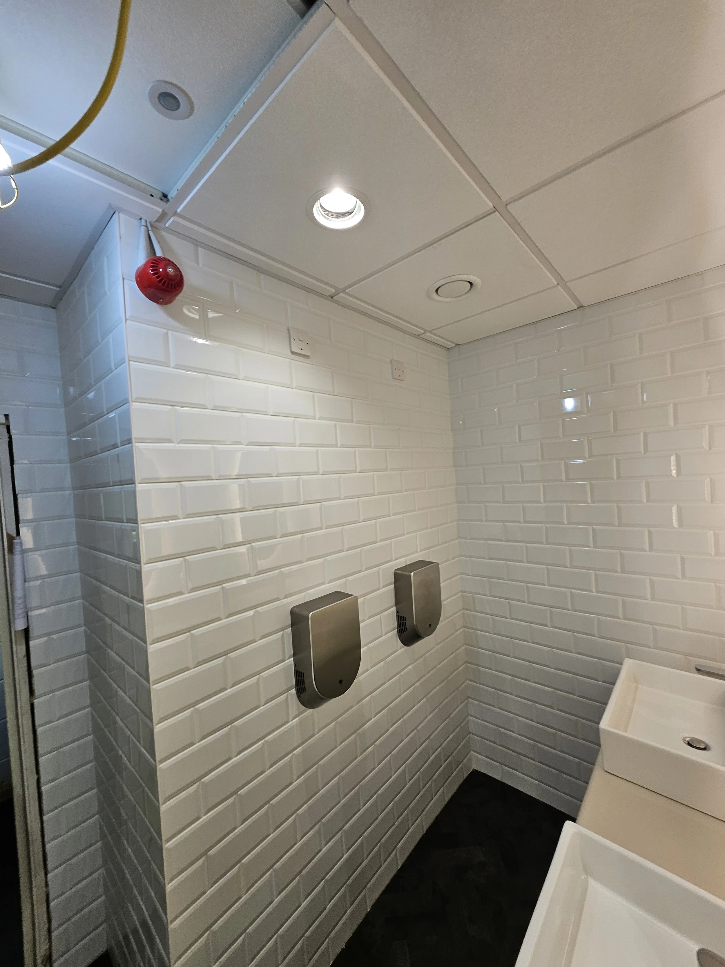 Public restroom with white subway tile walls, two hand dryers on the wall, and white sinks with metal faucets.