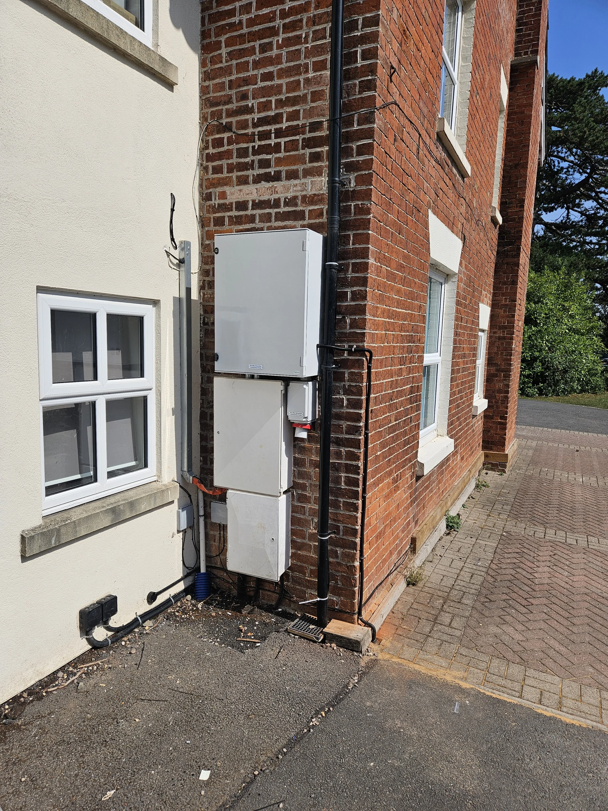 Exterior corner of a building with electrical boxes and wiring mounted on the wall, with a driveway and sidewalk nearby.