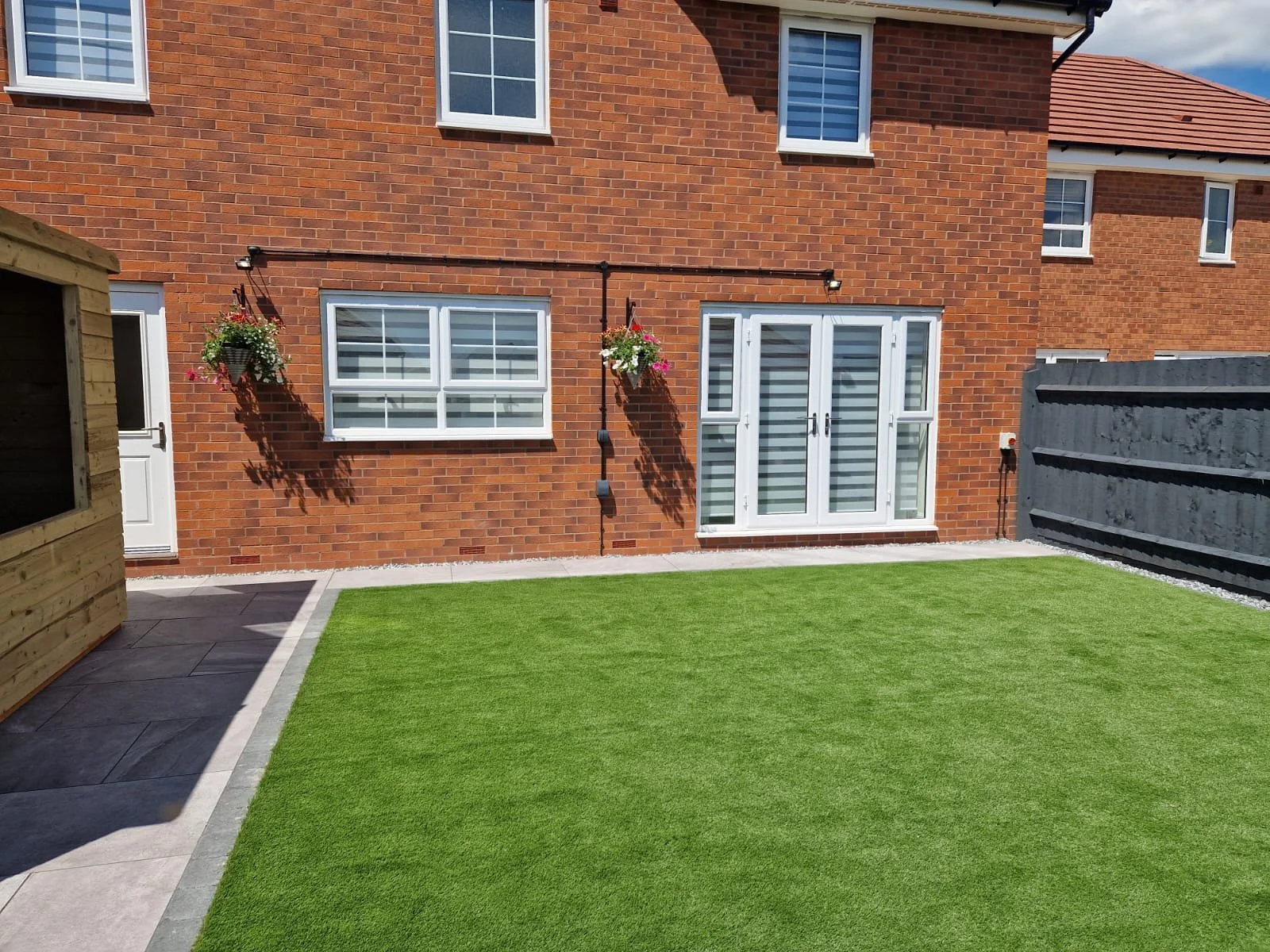 Backyard with green artificial grass, brick house with white-framed windows and doors, hanging flower baskets, fenced boundary, and a garden shed.