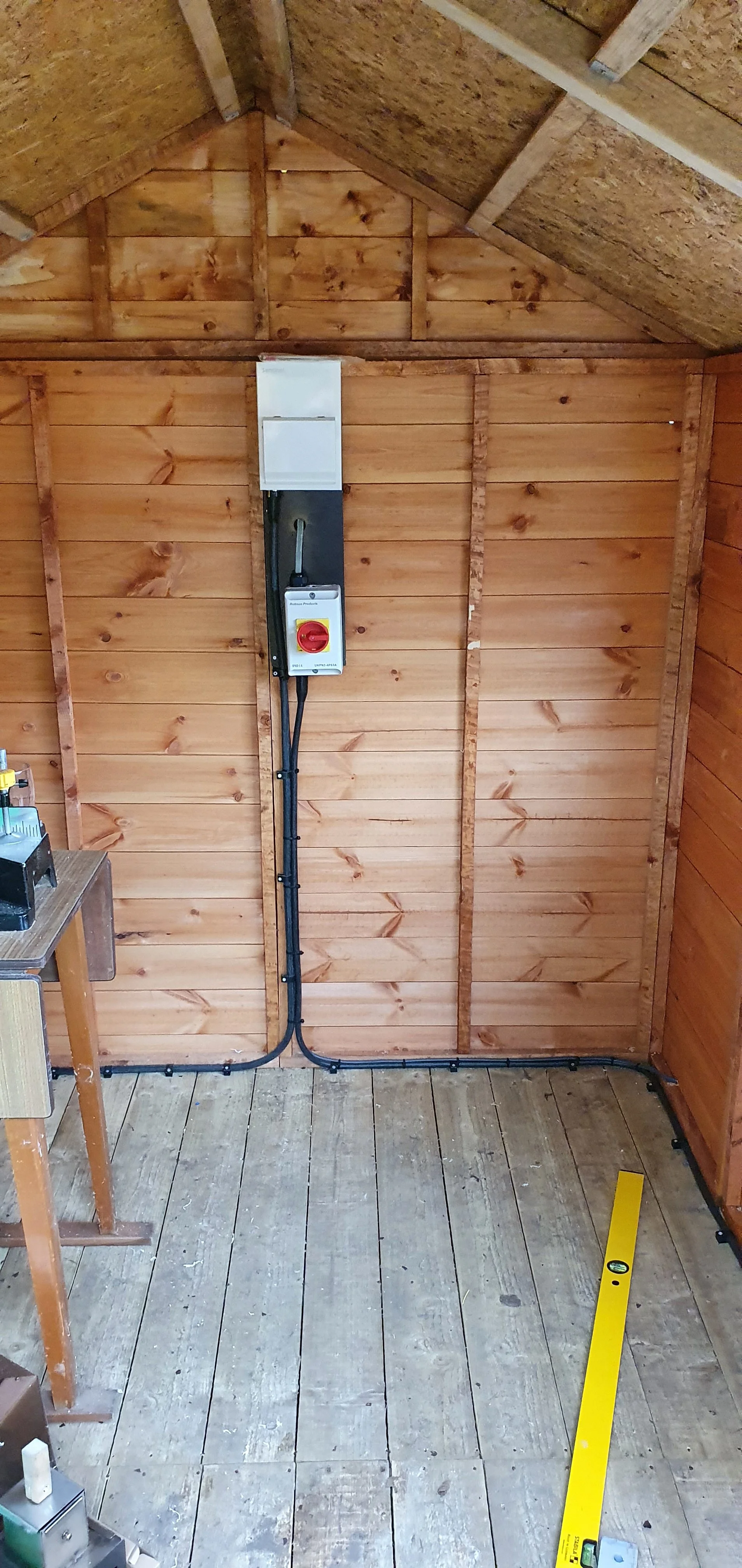 Interior of a wooden shed with exposed walls and ceiling. An electrical panel and wiring are mounted on the wall. The floor is unfinished wood with a yellow level tool resting on it.