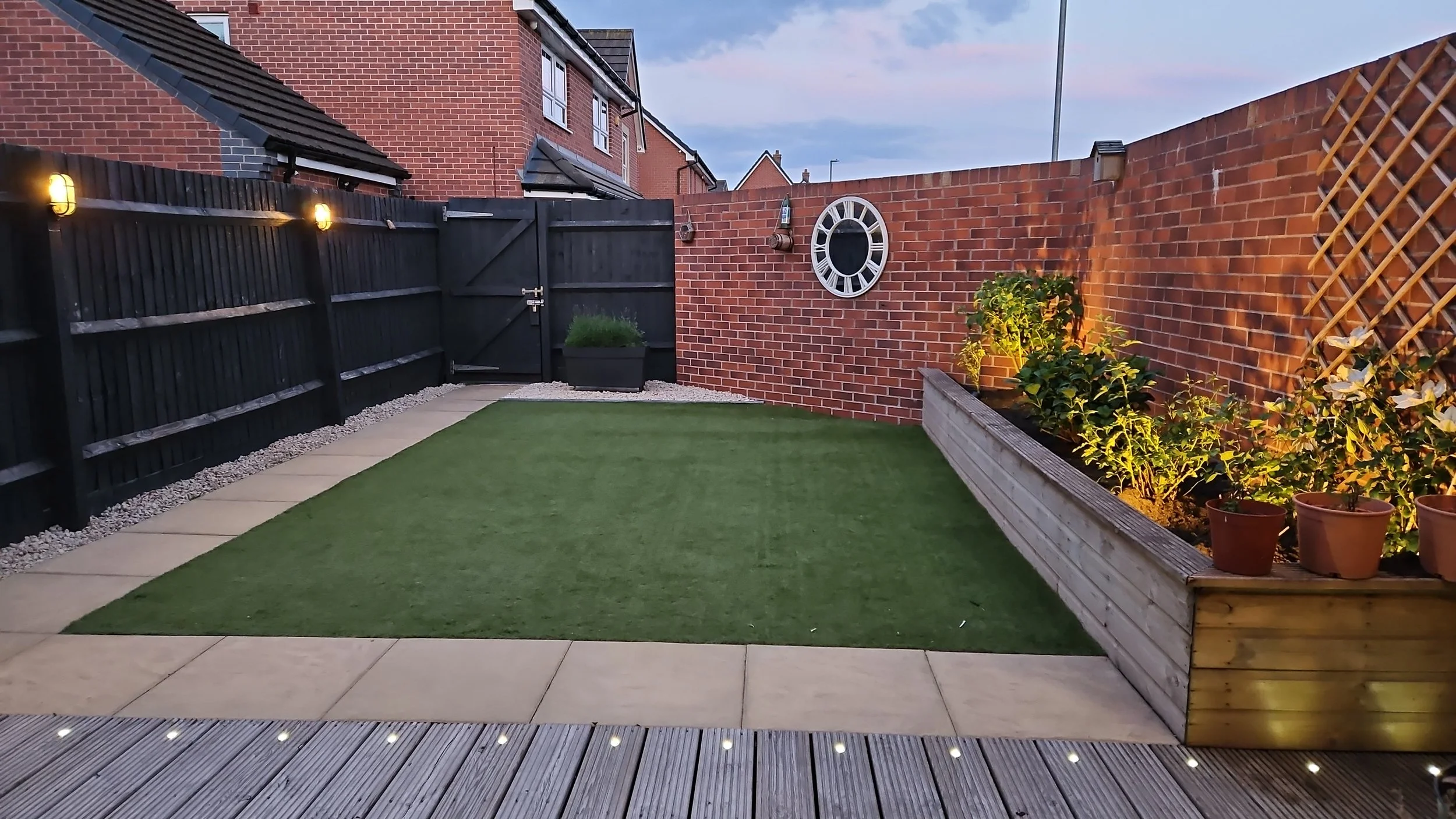 A small backyard with a green lawn, a paved border, a black fence with lights, a brick wall with a clock, a gate, and a garden bed with plants and potted flowers.