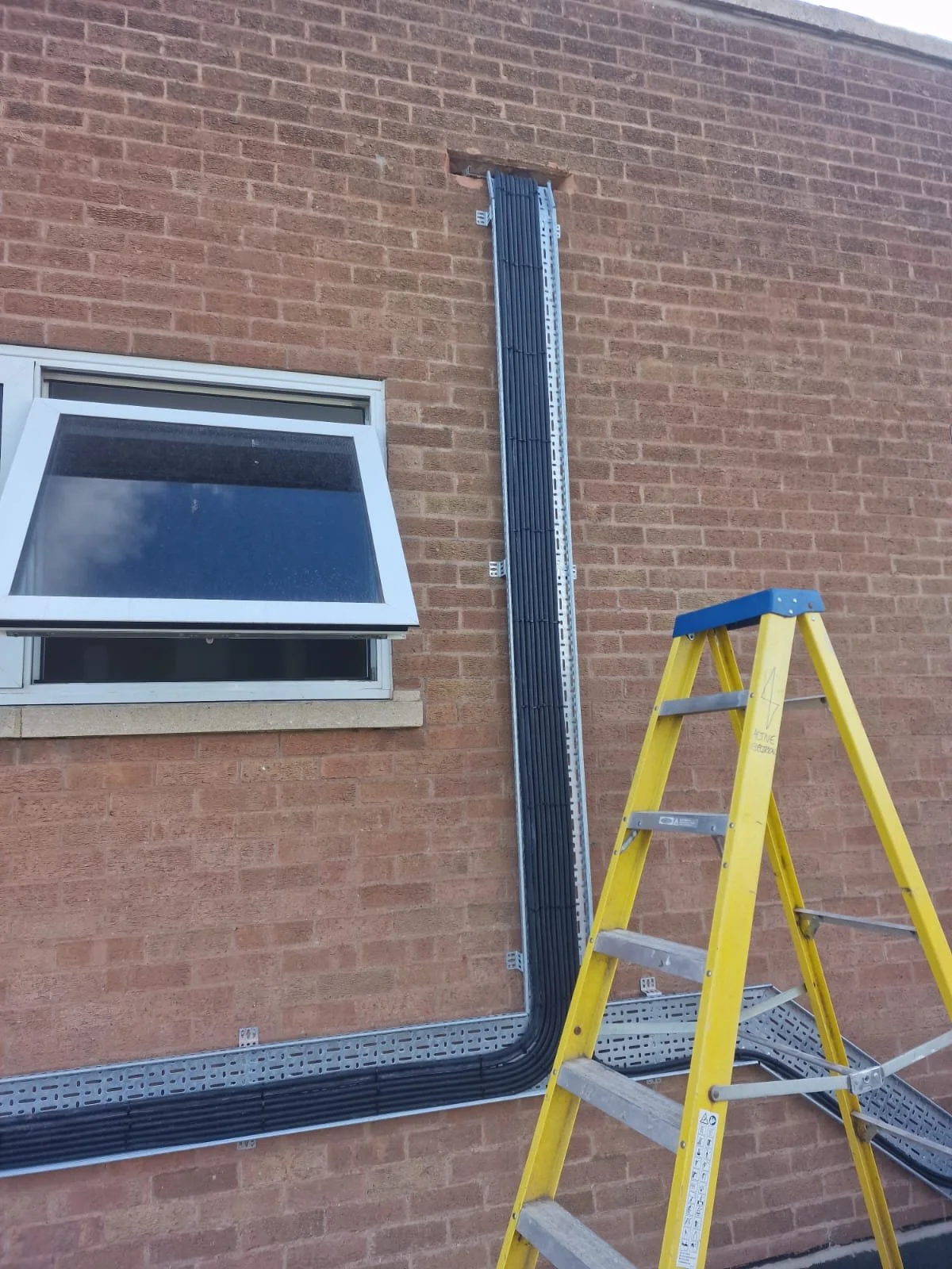 Electrical wiring and conduit installation on a brick building exterior near a window, with a yellow step ladder nearby.