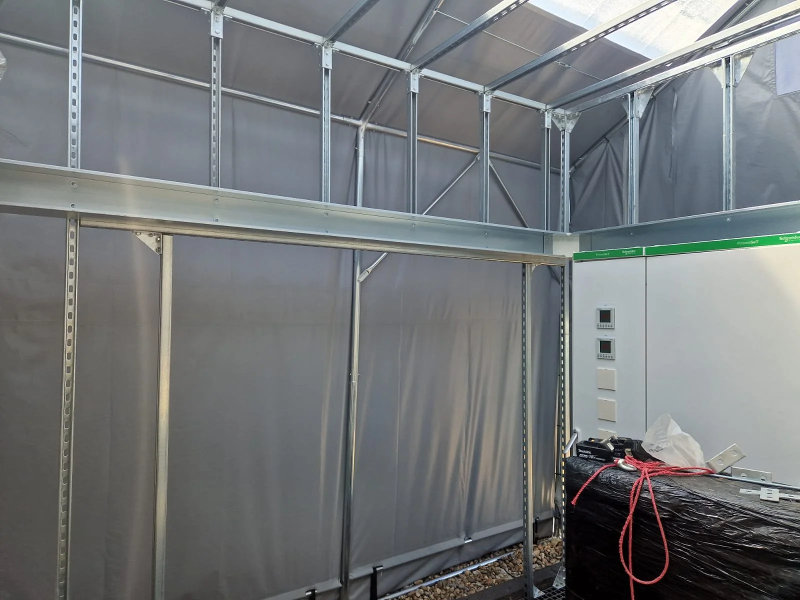 Interior of a metal construction site or temporary structure with gray walls, some electrical control panels, and construction tools and materials on a black wrapped pallet.