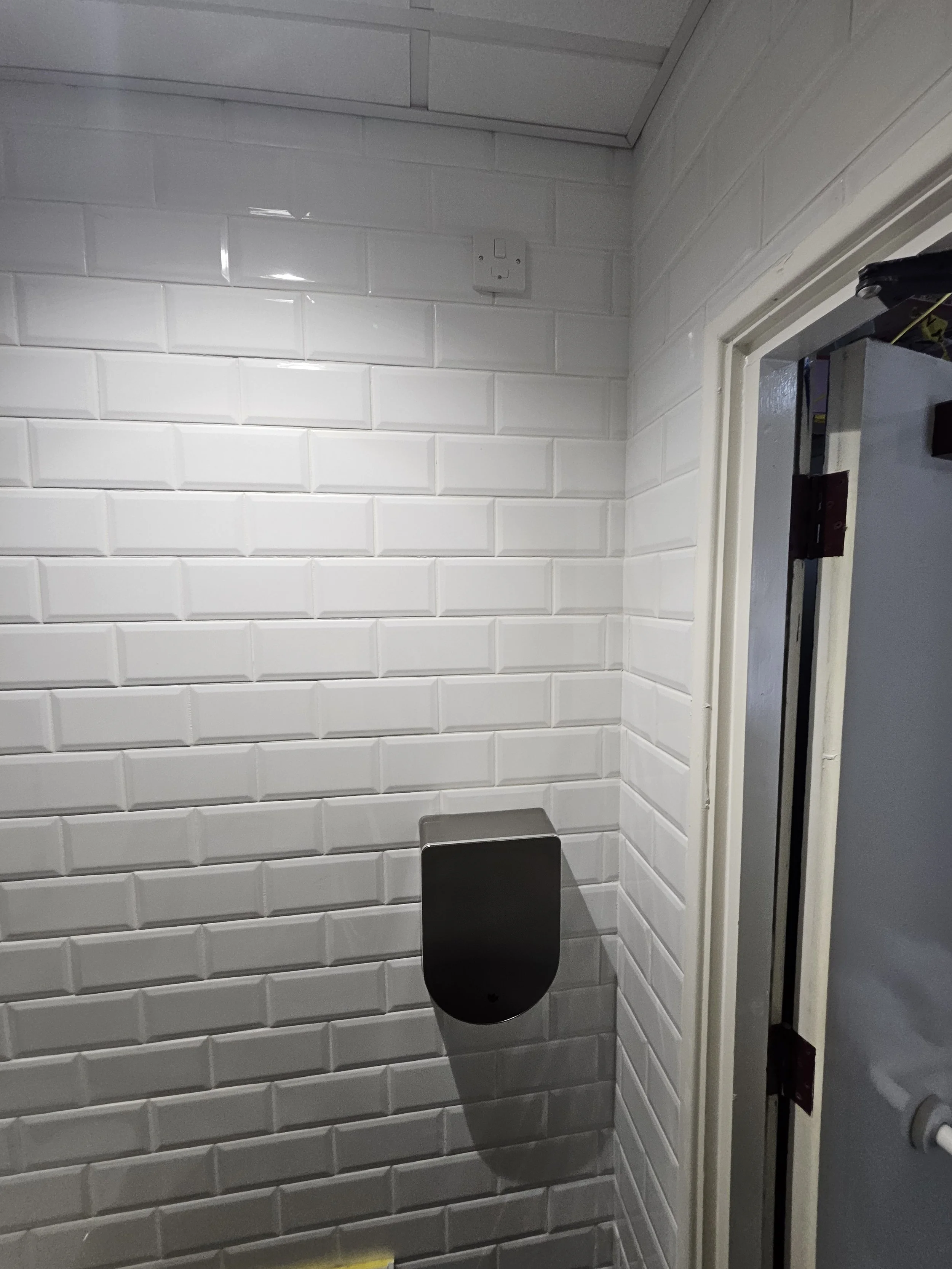 A white tiled wall with a black hand sanitizer dispenser and an electrical outlet near the ceiling, next to a doorway with a door frame.