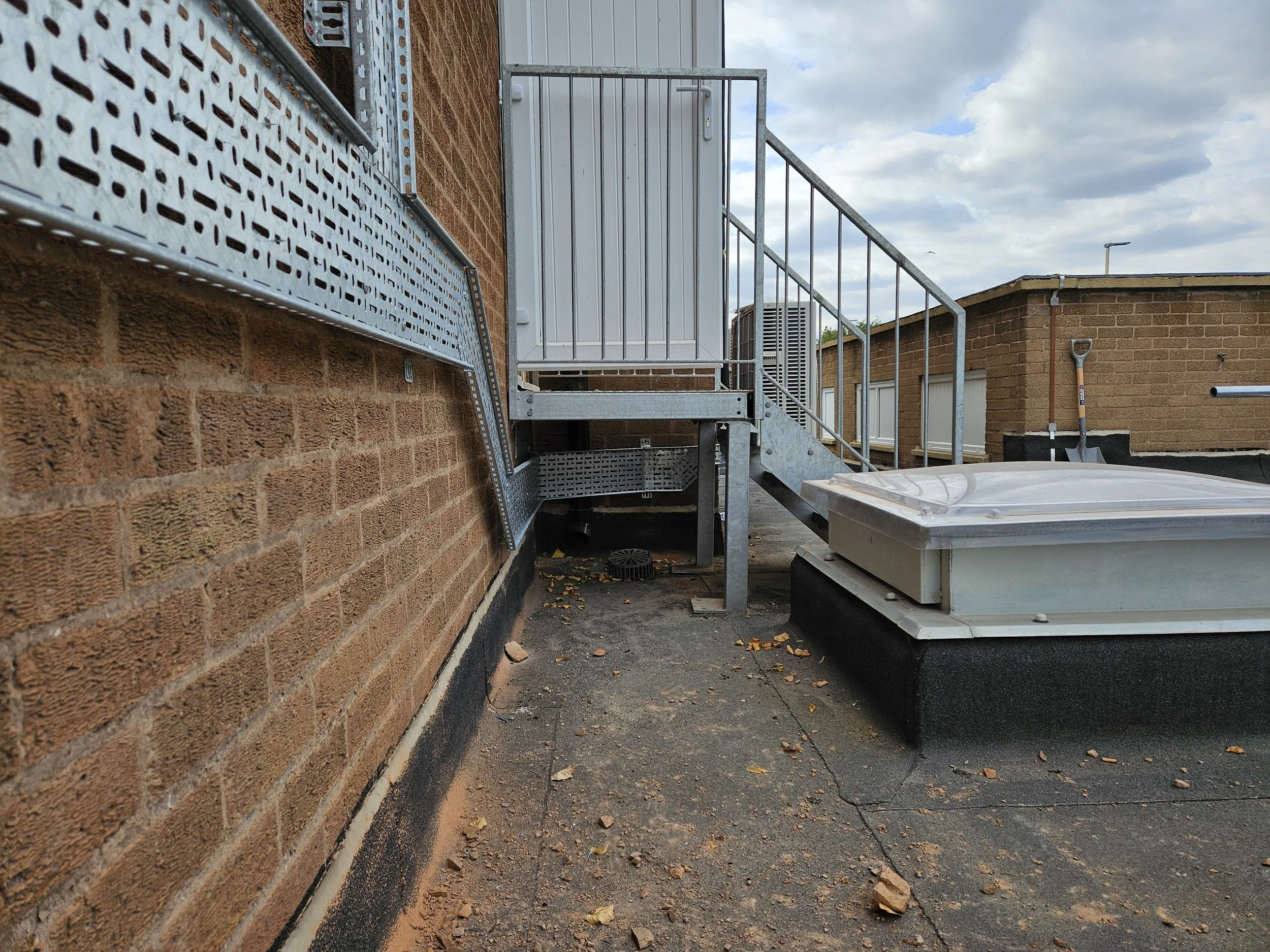 A rooftop view showing a brick wall with metal staircase, metal scaffolding, a white rooftop ventilation unit, and scattered leaves on the asphalt surface under a cloudy sky.