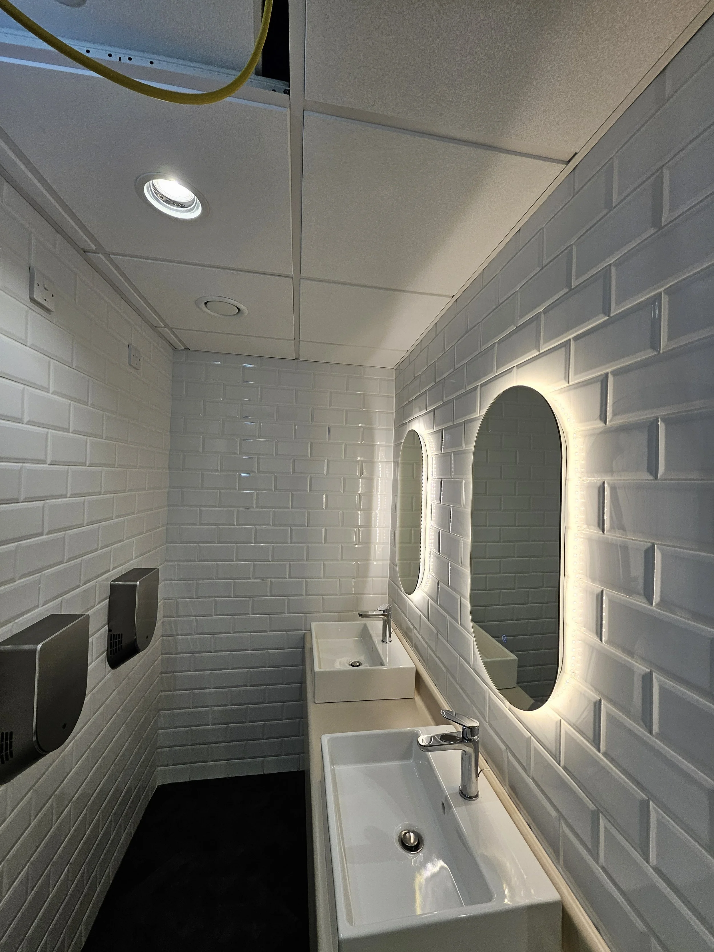A modern public restroom with white brick walls, two rectangular sinks with chrome faucets, two illuminated oval mirrors, and two stainless steel hand dryers on the wall.
