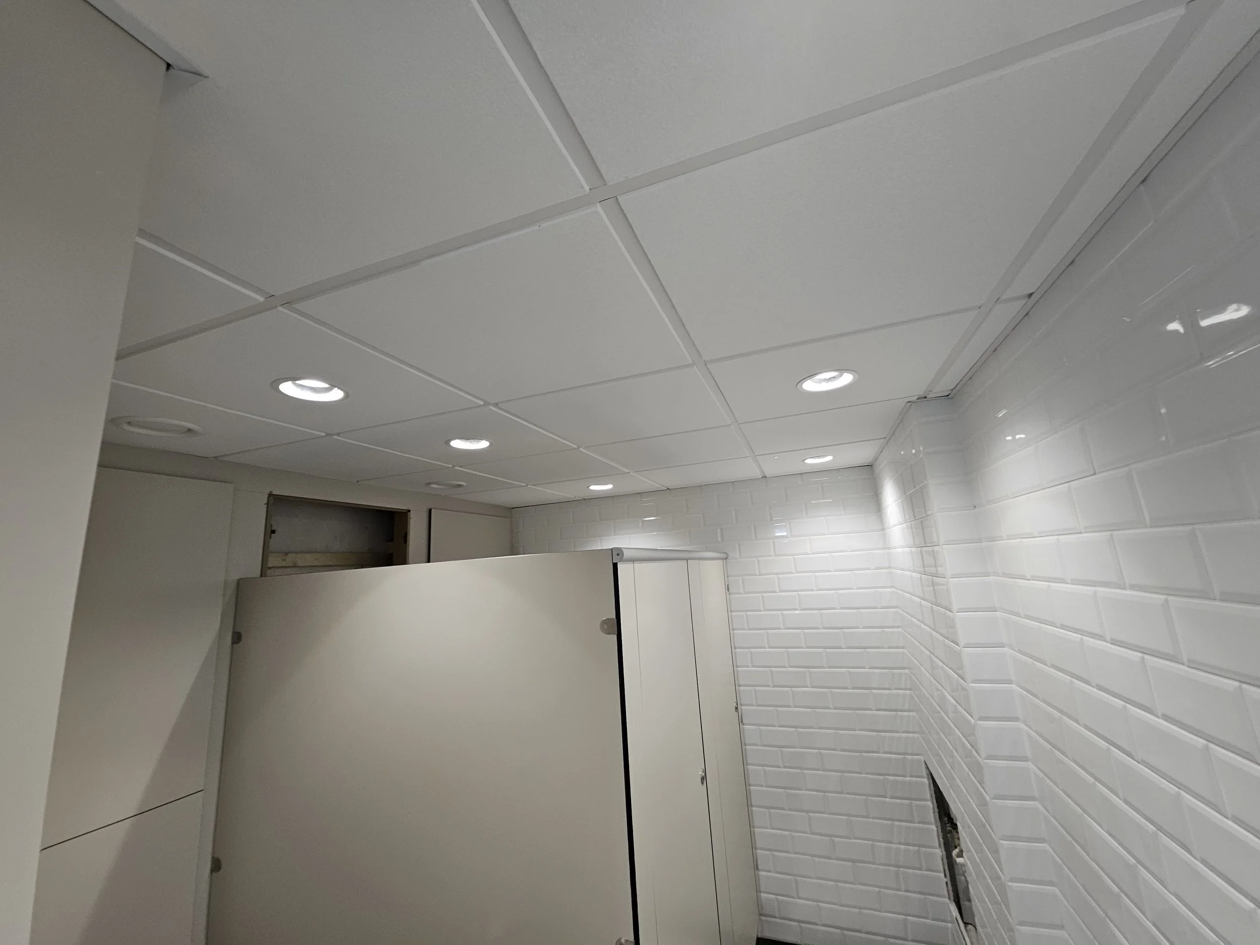 A white tiled bathroom wall with an opening in the wall, ceiling with recessed lighting, and bathroom stall walls.