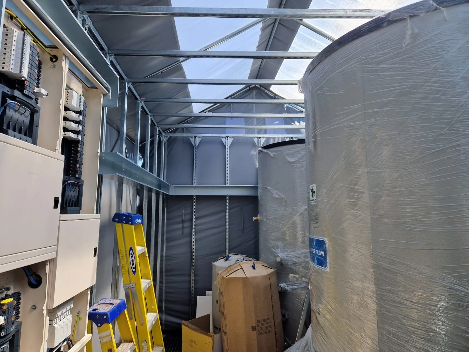 The interior of a construction or industrial space with exposed metal framing, electrical panels, a yellow step ladder, and a large wrapped cylindrical tank.