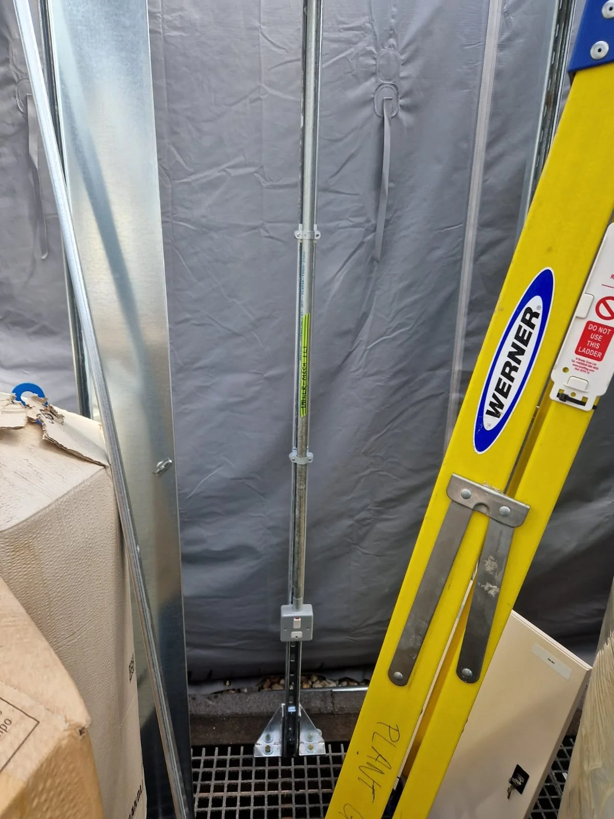 A construction workspace with a metal pole, a yellow WERNER ladder, and gray temporary walls. The work area appears to be in progress.
