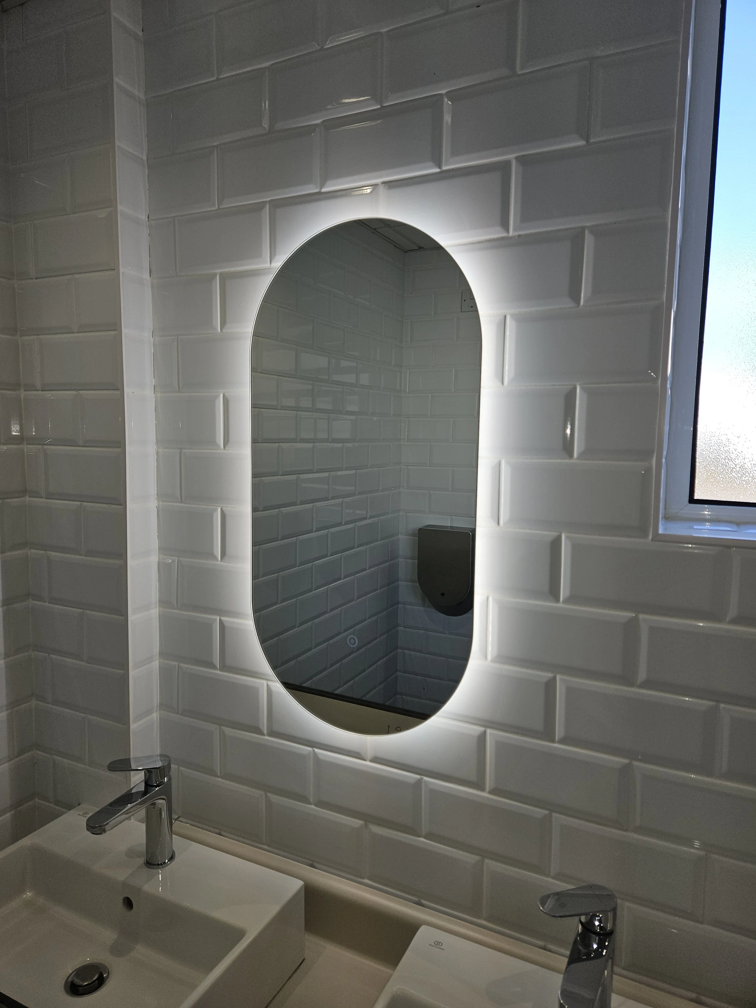 A bathroom with white subway tile walls, two sinks with chrome faucets, and an oval backlit mirror. There is a window on the right side.