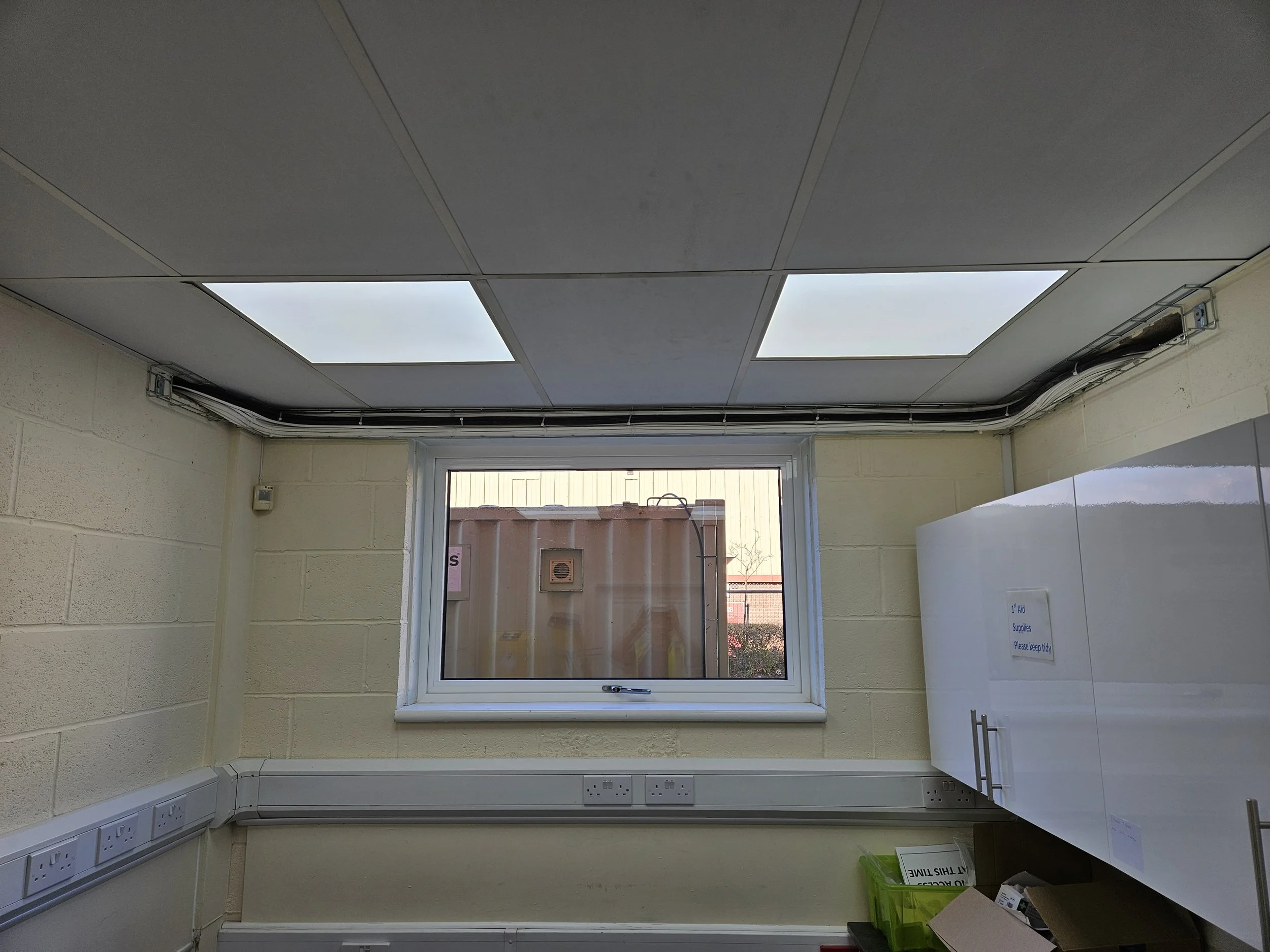 Interior view of a small room with a window showing an outdoor container and fence, ceiling with recessed lighting panels, cream walls, and electrical outlets along the bottom of the walls.