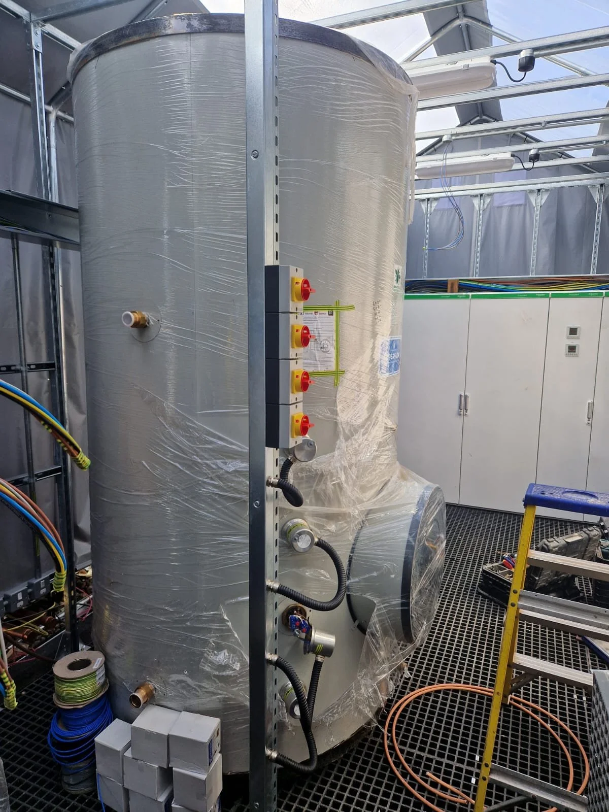 A large cylindrical industrial tank wrapped in plastic, installed in a metal-framed greenhouse with electrical wires and tools nearby.