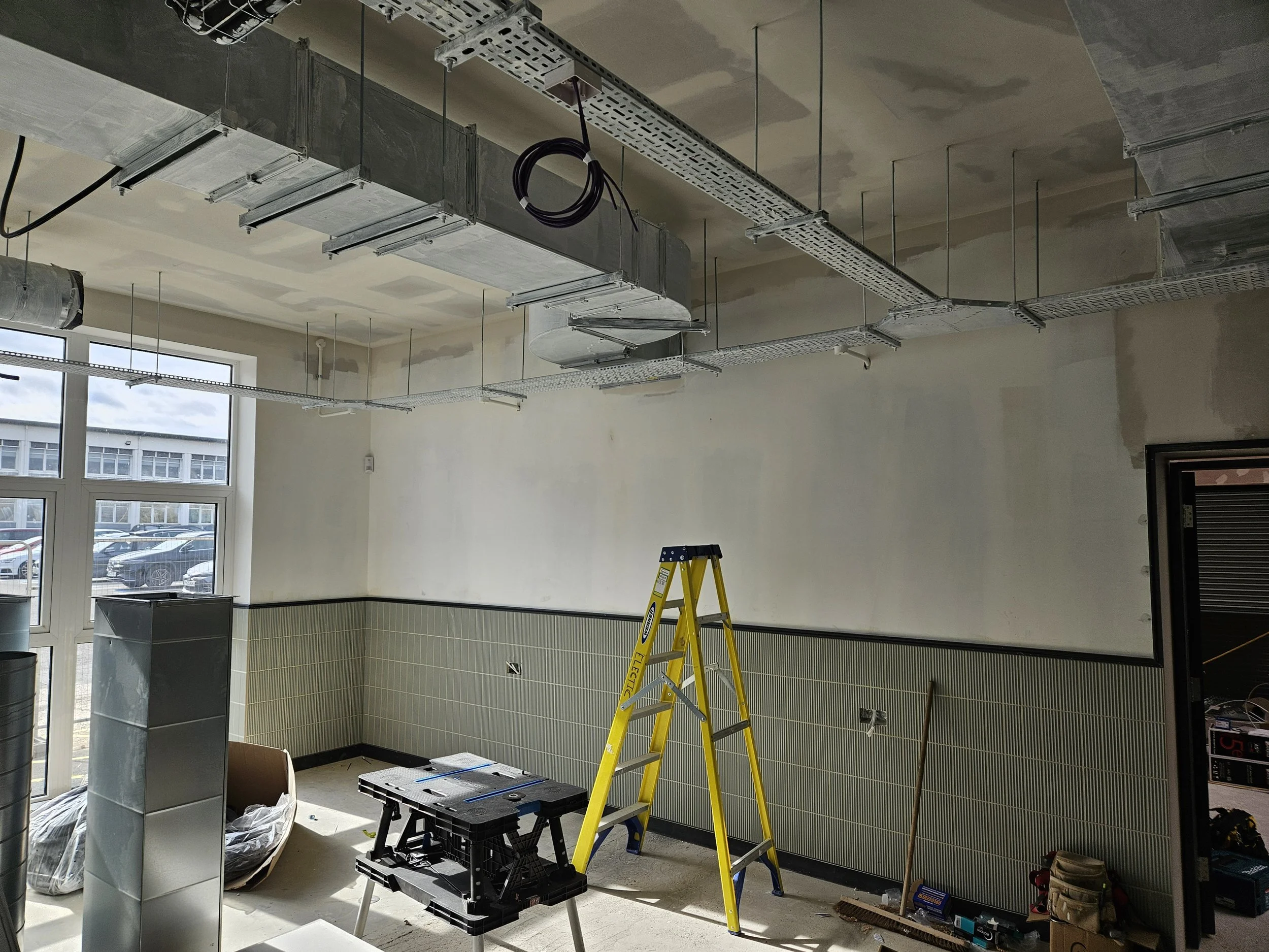 Construction site interior with a yellow ladder, a black worktable, construction tools, and partially finished walls and ceilings, with large windows showing parked cars outside.