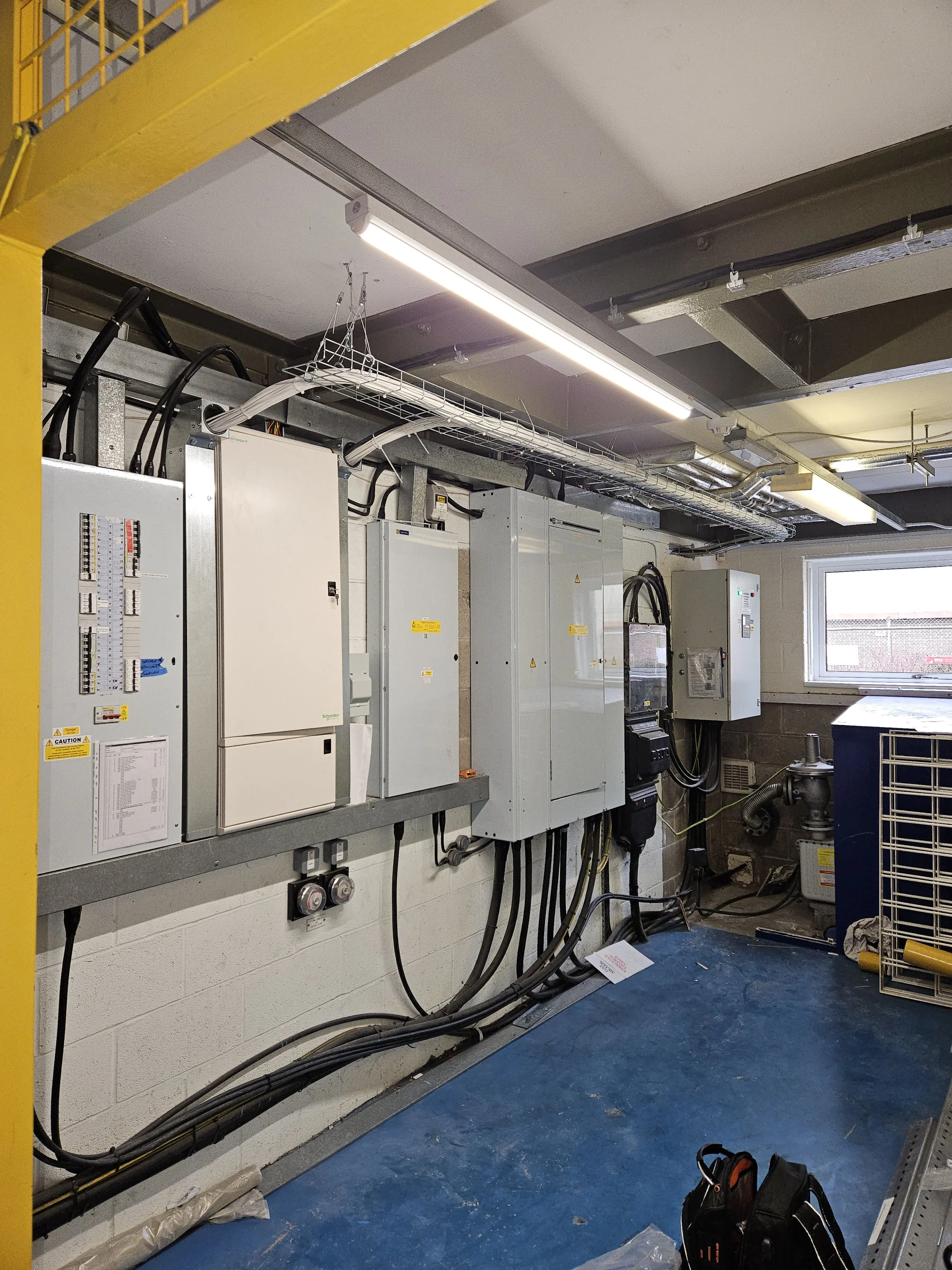 An industrial electrical room with multiple control panels, circuit breakers, and wiring, illuminated by overhead fluorescent lighting, with a window on the right side and various tools and equipment on the floor.