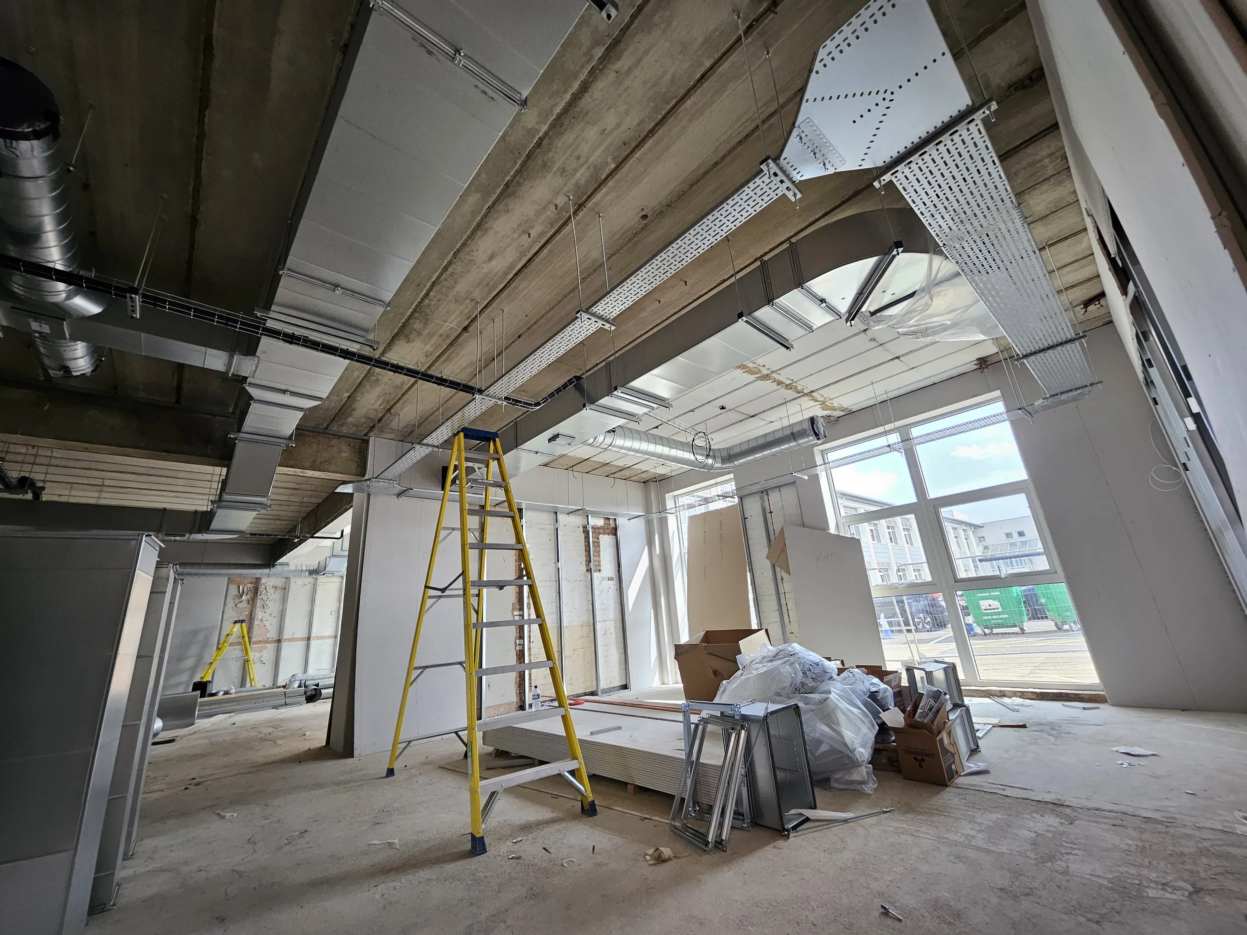 Construction site with ladders, building materials, and installation in progress, large windows letting in sunlight.
