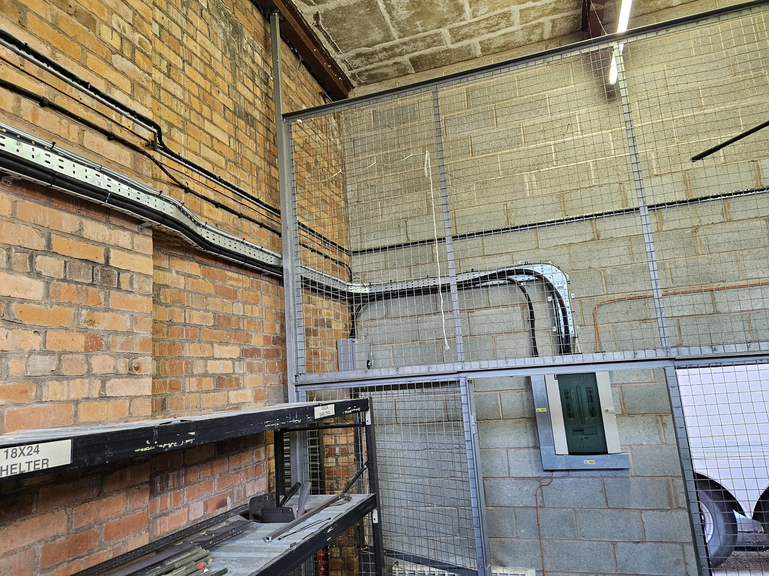 Industrial storage area with brick and cinder block walls, metal wiring channels, metal grid fencing, electrical components, and a partially visible washing machine.