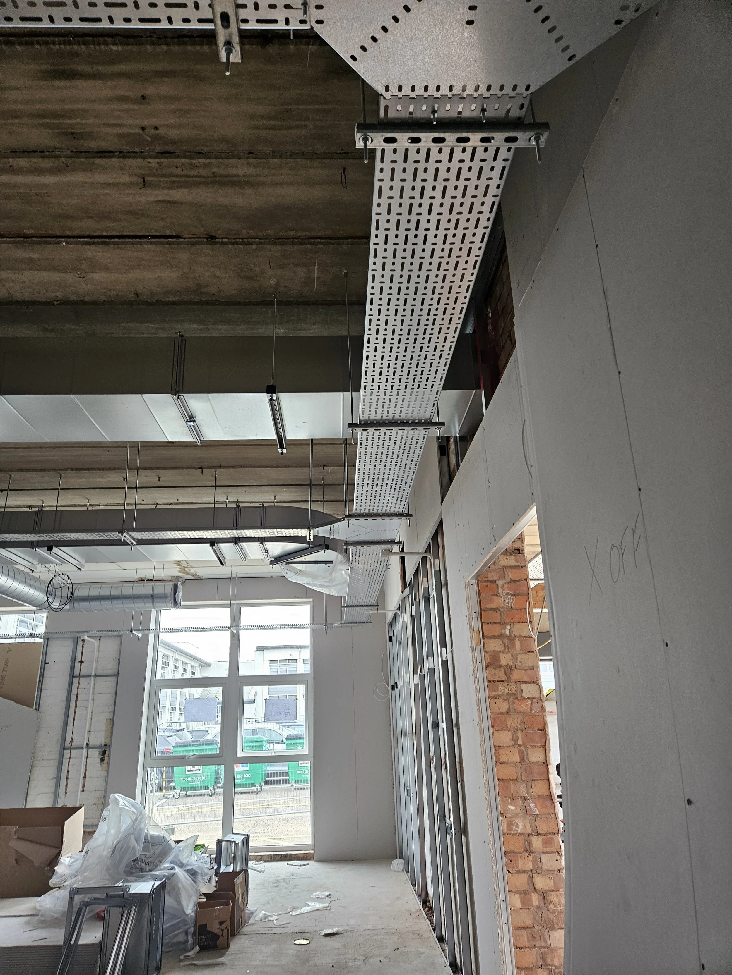 Interior of a building under construction with metal ceiling panels, exposed brick wall, and large window. Construction materials and debris are scattered on the floor.