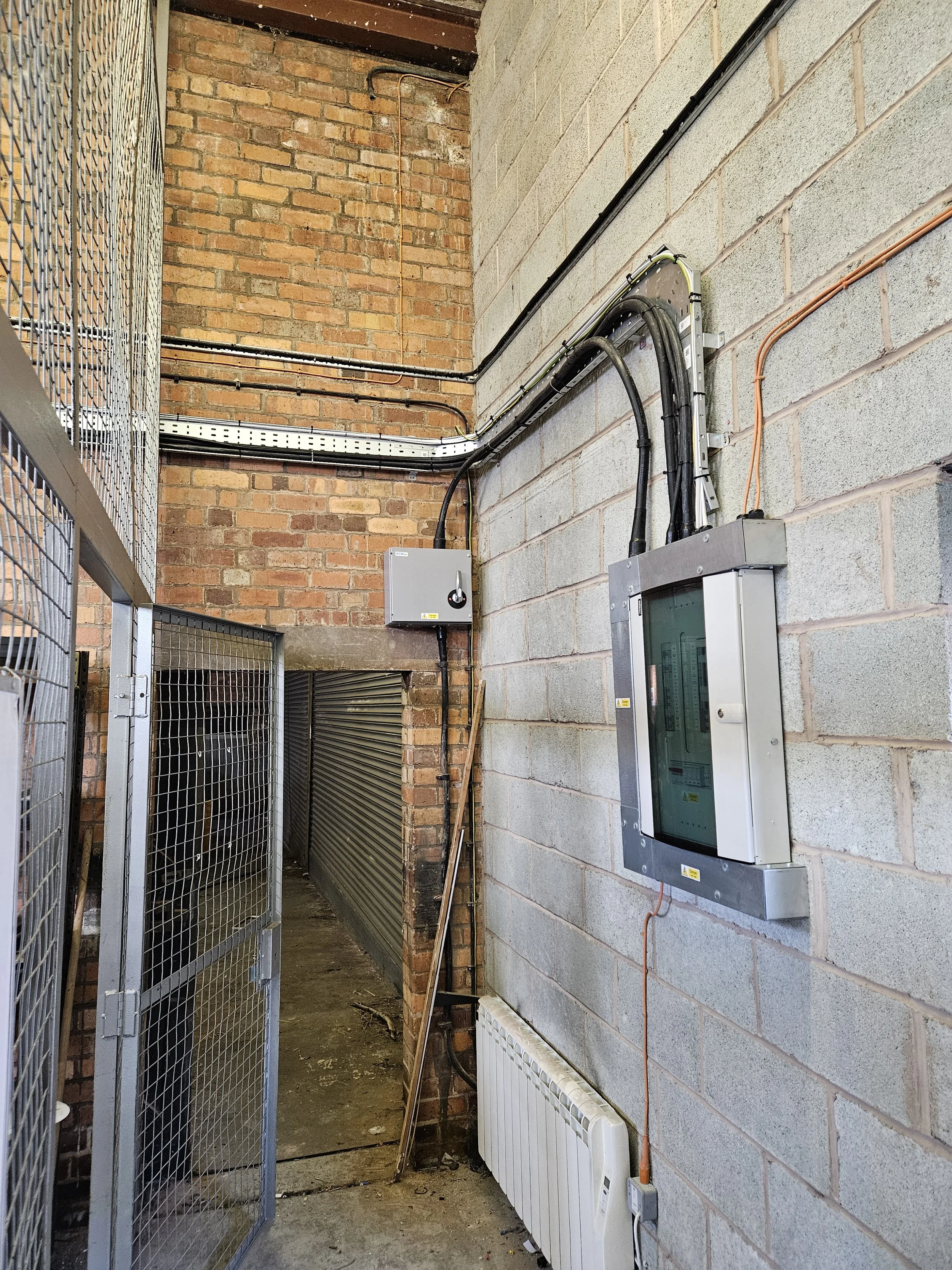 Electrical control panel and wiring on cinder block wall, with a radiator below and a metal gate on the left.