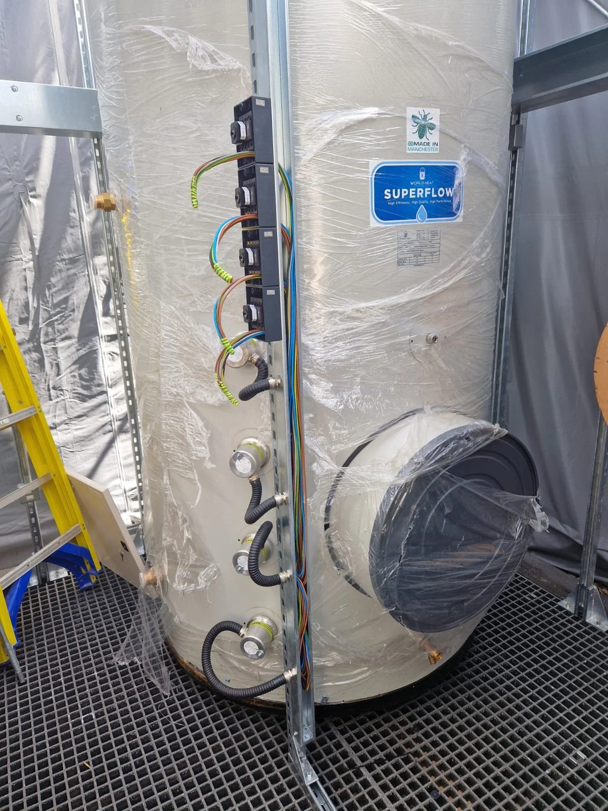 Large industrial machine wrapped in plastic with labeled stickers, wires connected in the middle, and a black exhaust port on the right side. Yellow ladder on the left and metal framework around.