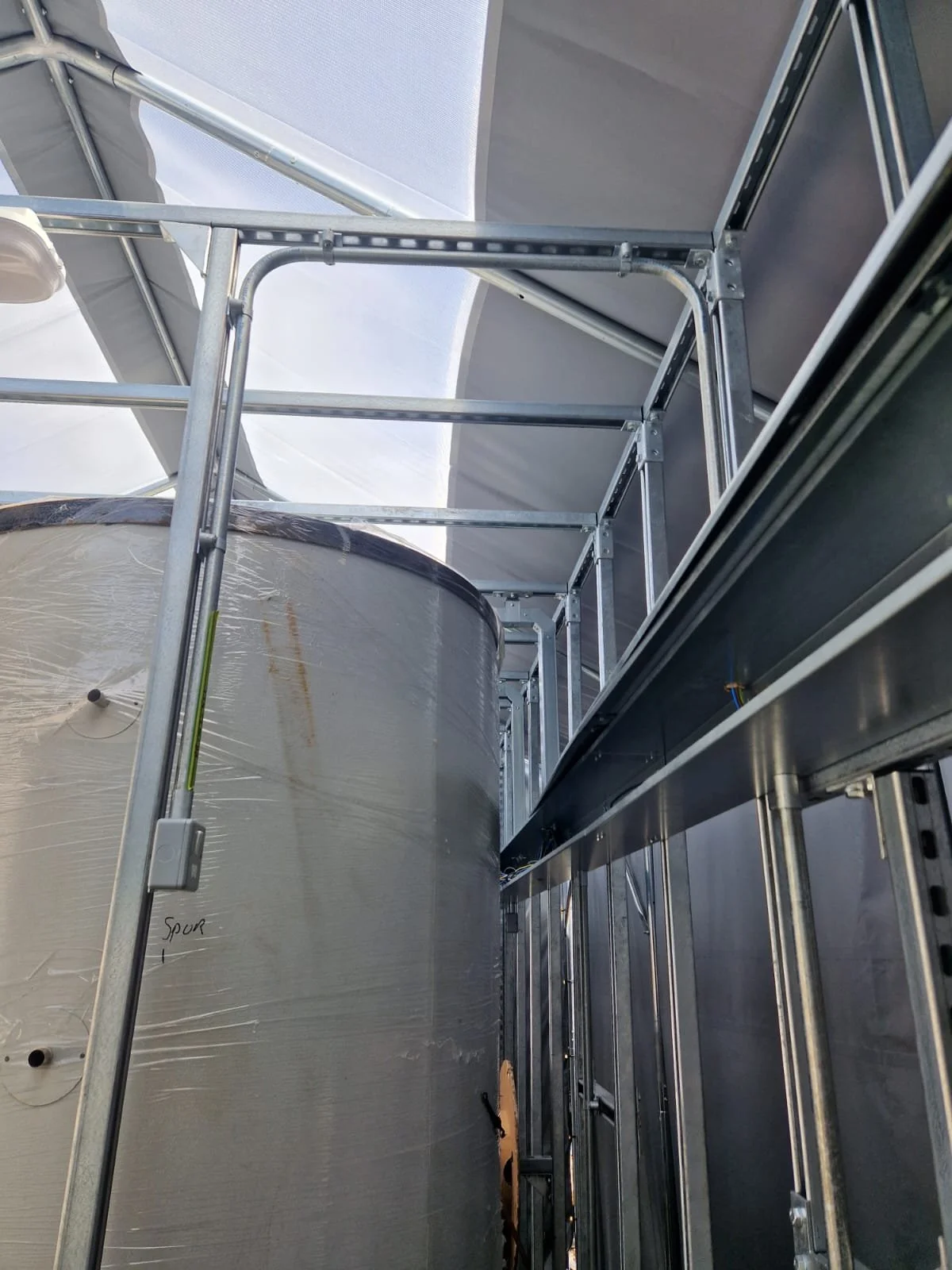 Inside a greenhouse, metal scaffolding surrounds a large cylindrical water tank covered with plastic wrap, with a part of the sky visible through the transparent ceiling.