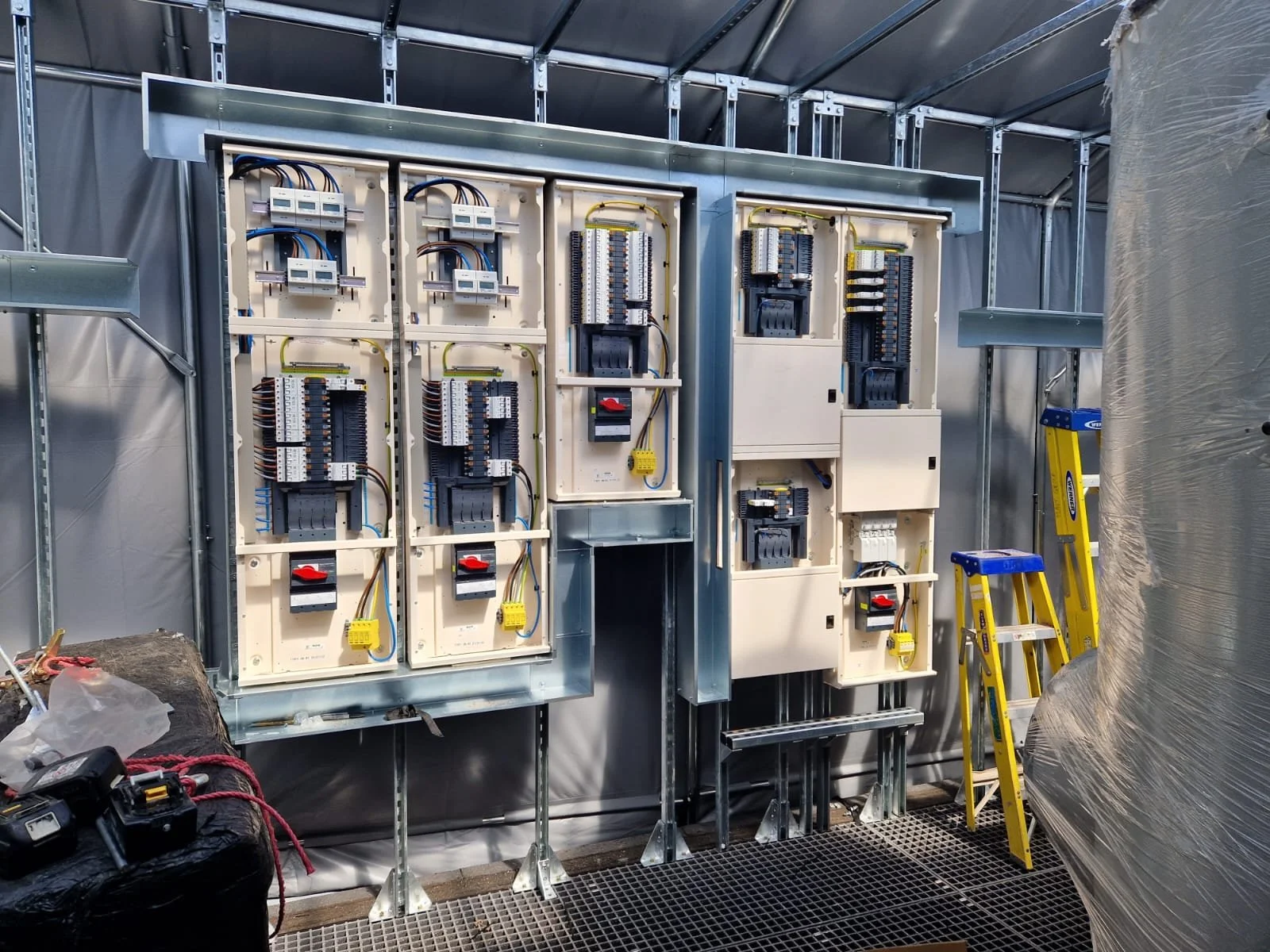 Interior view of electrical control panels with various switches, wiring, and circuit breakers installed. Ladder and construction materials on site.