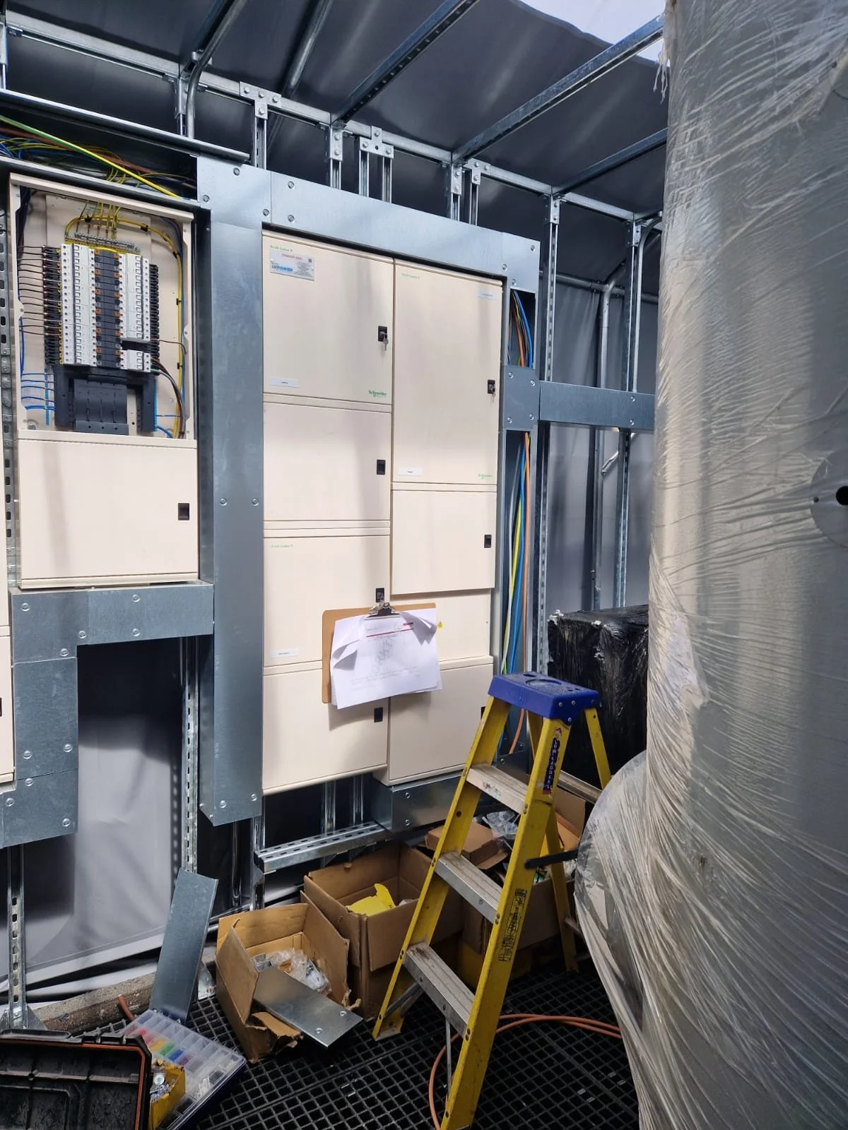 Electrical panel mounted on a metal frame inside a utility room, with a yellow step ladder, cardboard boxes, tools, and wiring around.