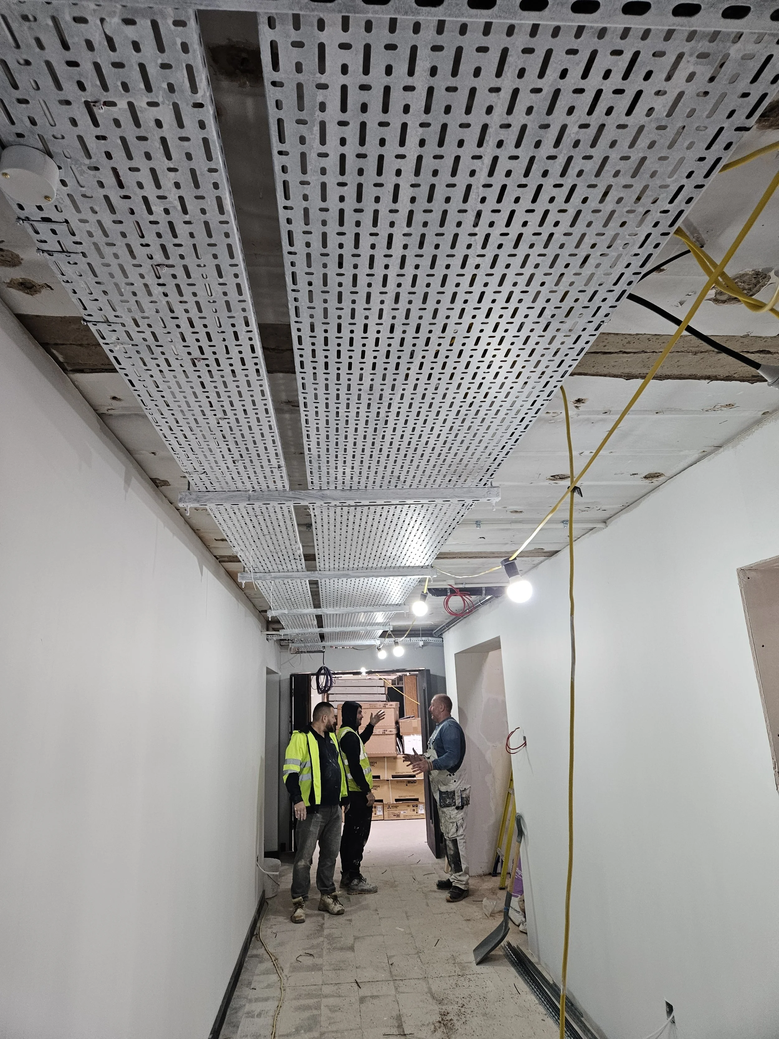 Construction workers in a hallway under renovation, with new ceiling panels and hanging lights, discussions taking place.