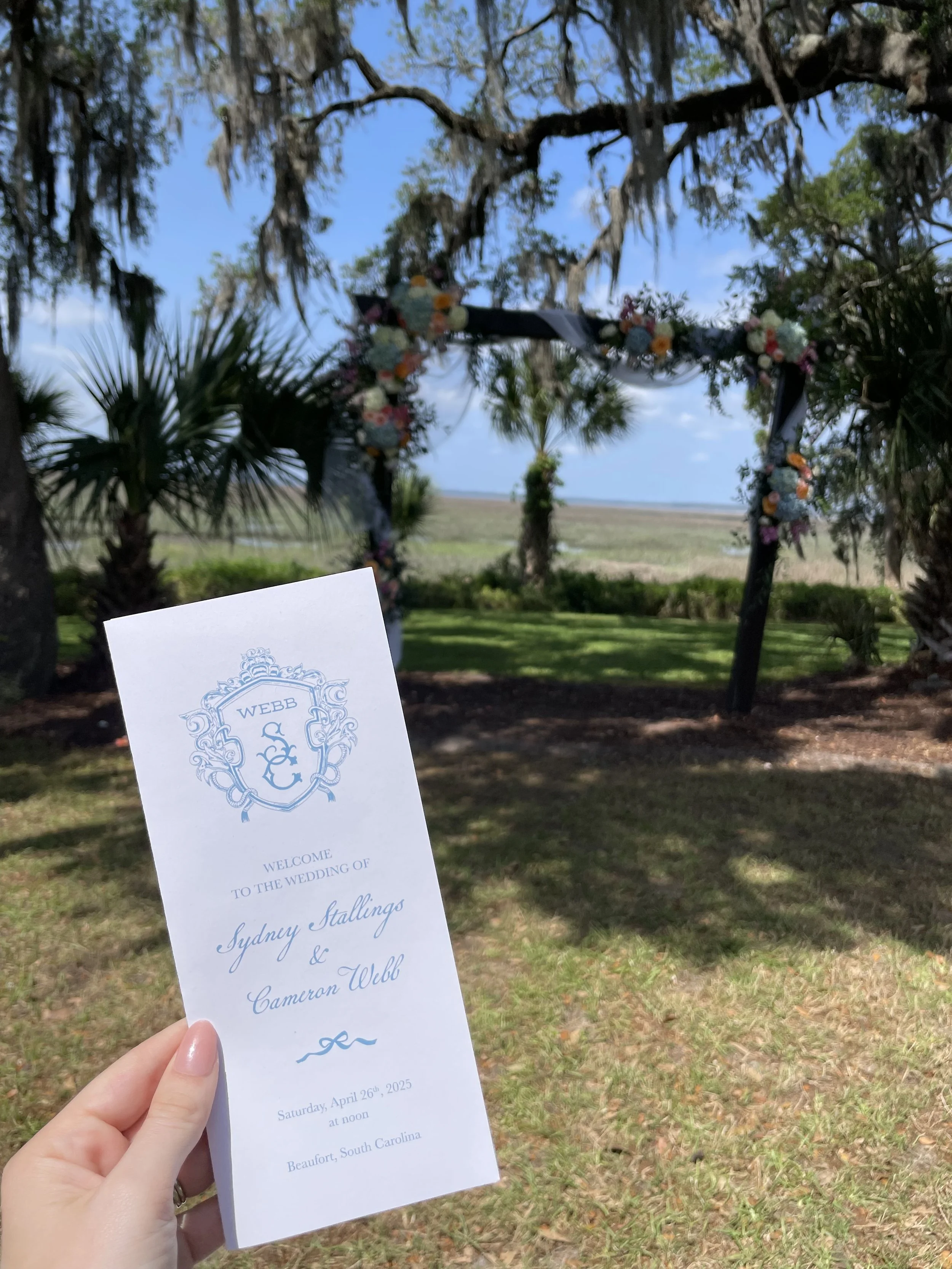 A wedding program is held outdoors in a green landscape with trees and a floral arch in the background. The program is titled welcoming a wedding of Sydney Stallings and Cameron Webb, scheduled for April 26, 2025, at noon in Beaufort, South Carolina.
