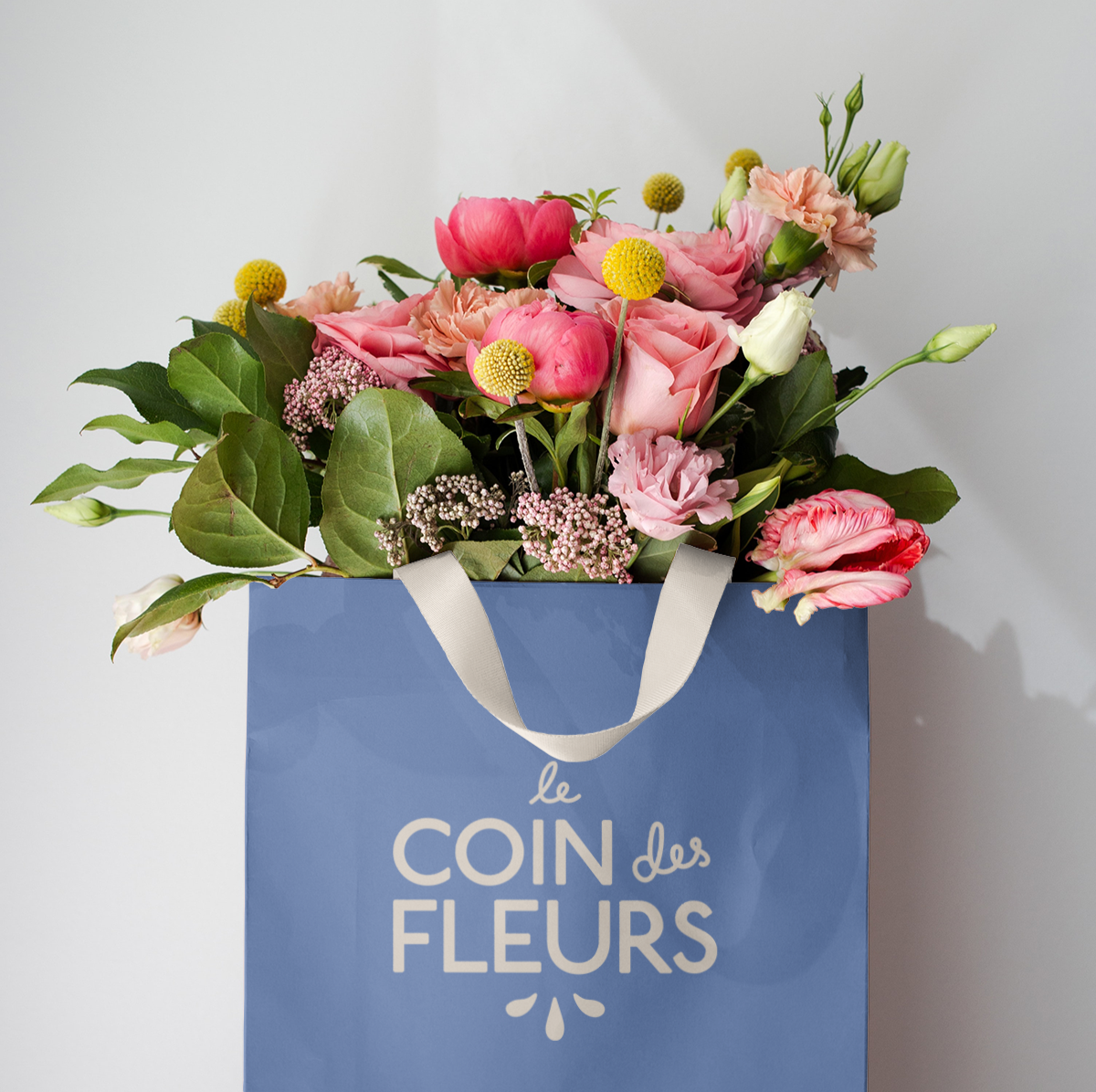 A bouquet of pink, white, and peach flowers in a blue shopping bag with white handles and the words "Le Coin des Fleurs" printed on it, against a plain light background.