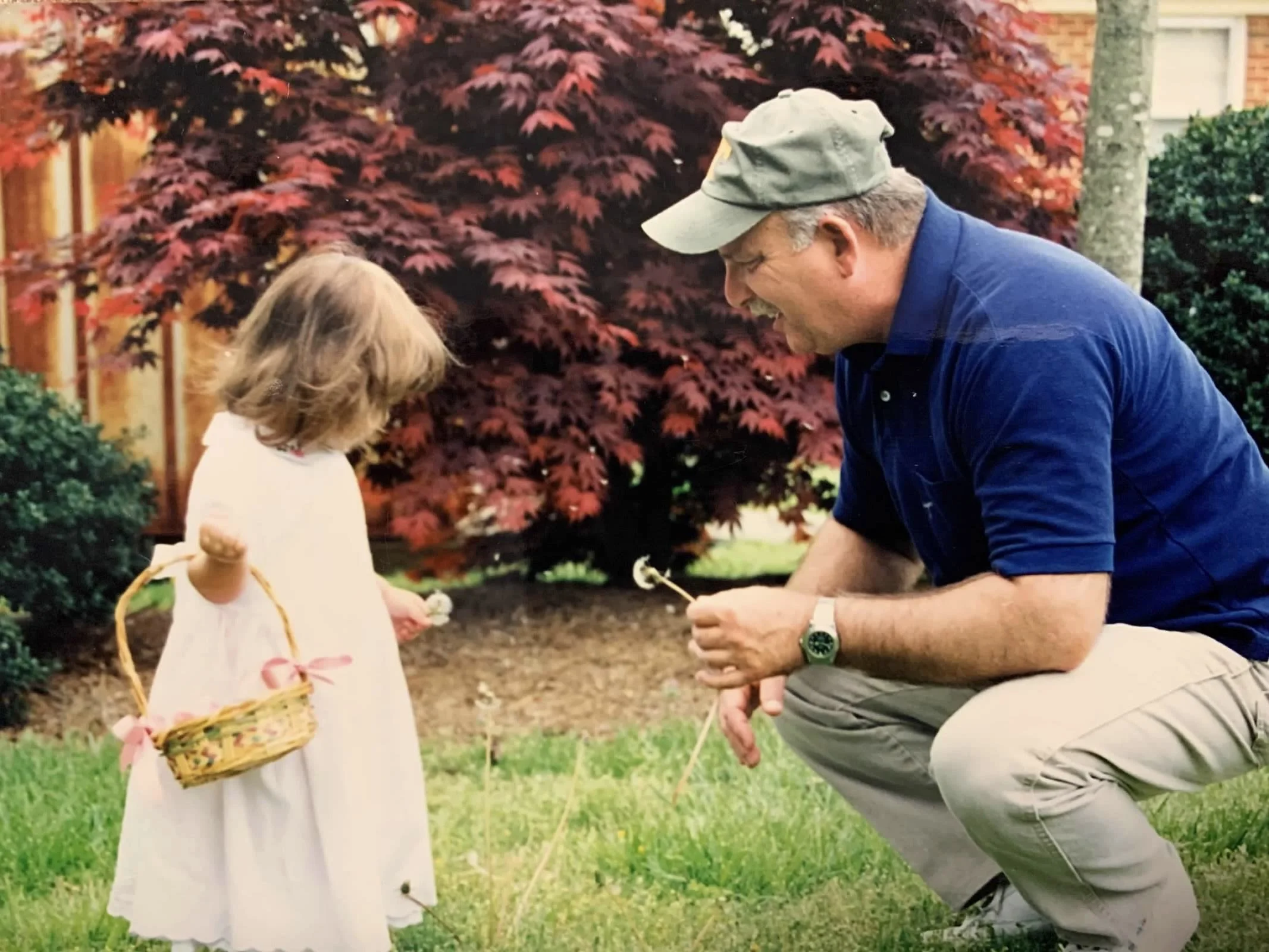 A man and young girl in a garden, with the man squatting and talking to the girl, who is holding a wicker basket with pink ribbons, and they are both holding small flowers or dandelions.