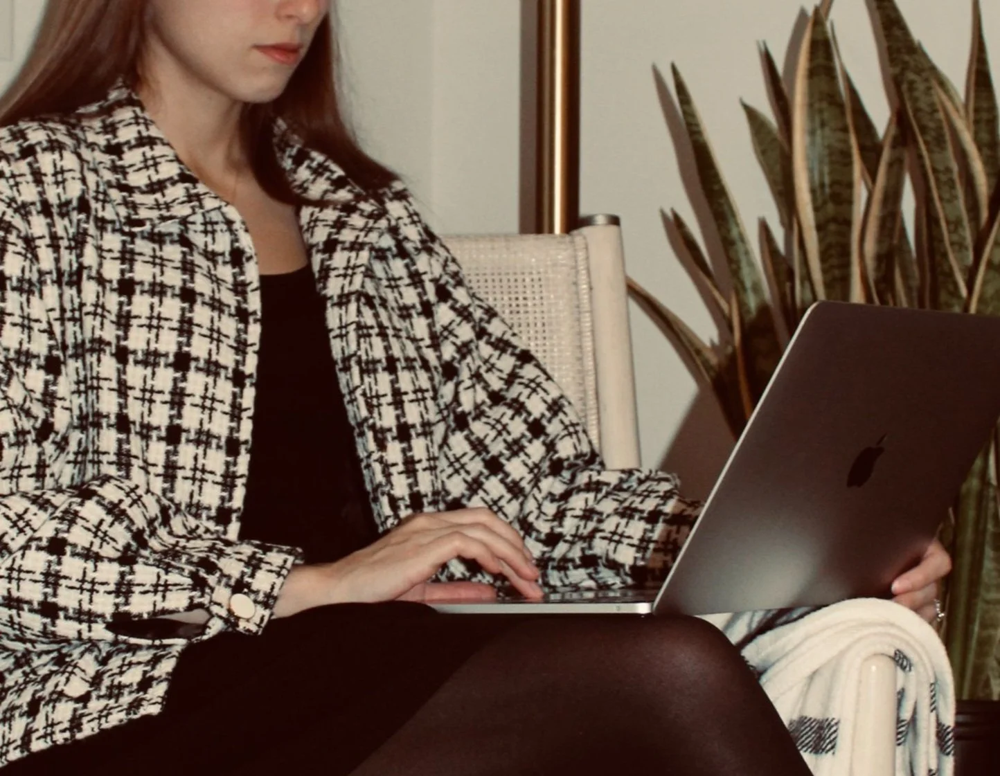 Woman with reddish hair wearing a black and white plaid blazer, sitting on a chair, using a laptop, with a large potted plant and a blanket nearby.