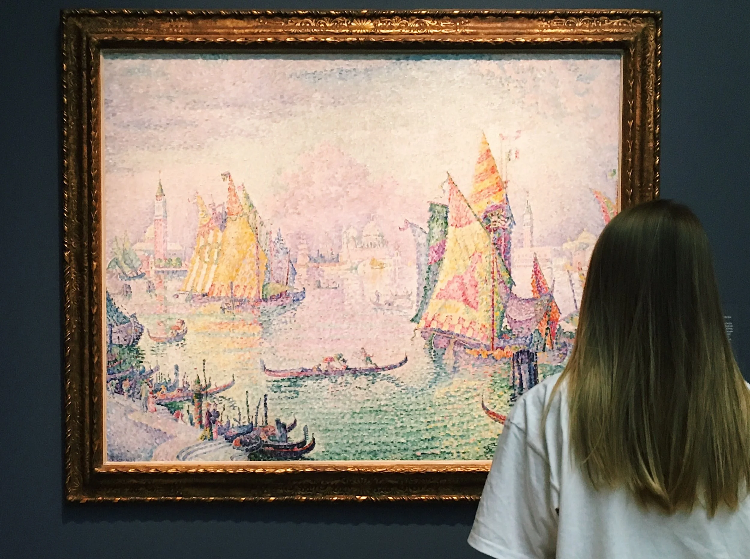 A person with long brown hair wearing a white shirt is viewing a painting of boats with colorful sails on a waterway, in a framed artwork in an art gallery.
