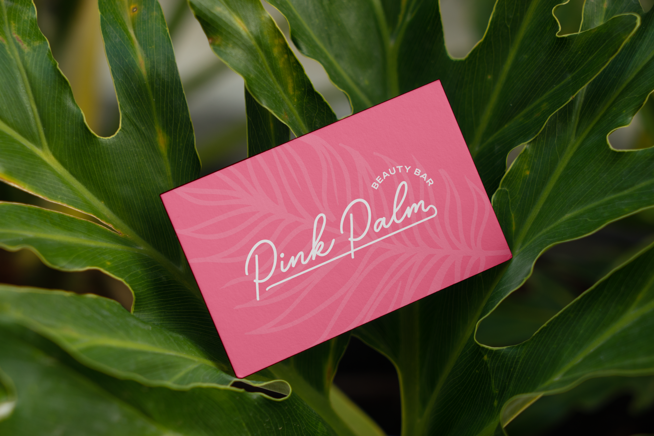 Pink business card with white text and leaf pattern, resting on large green tropical leaves. The card reads 'Pink Palm Beauty Bar'.