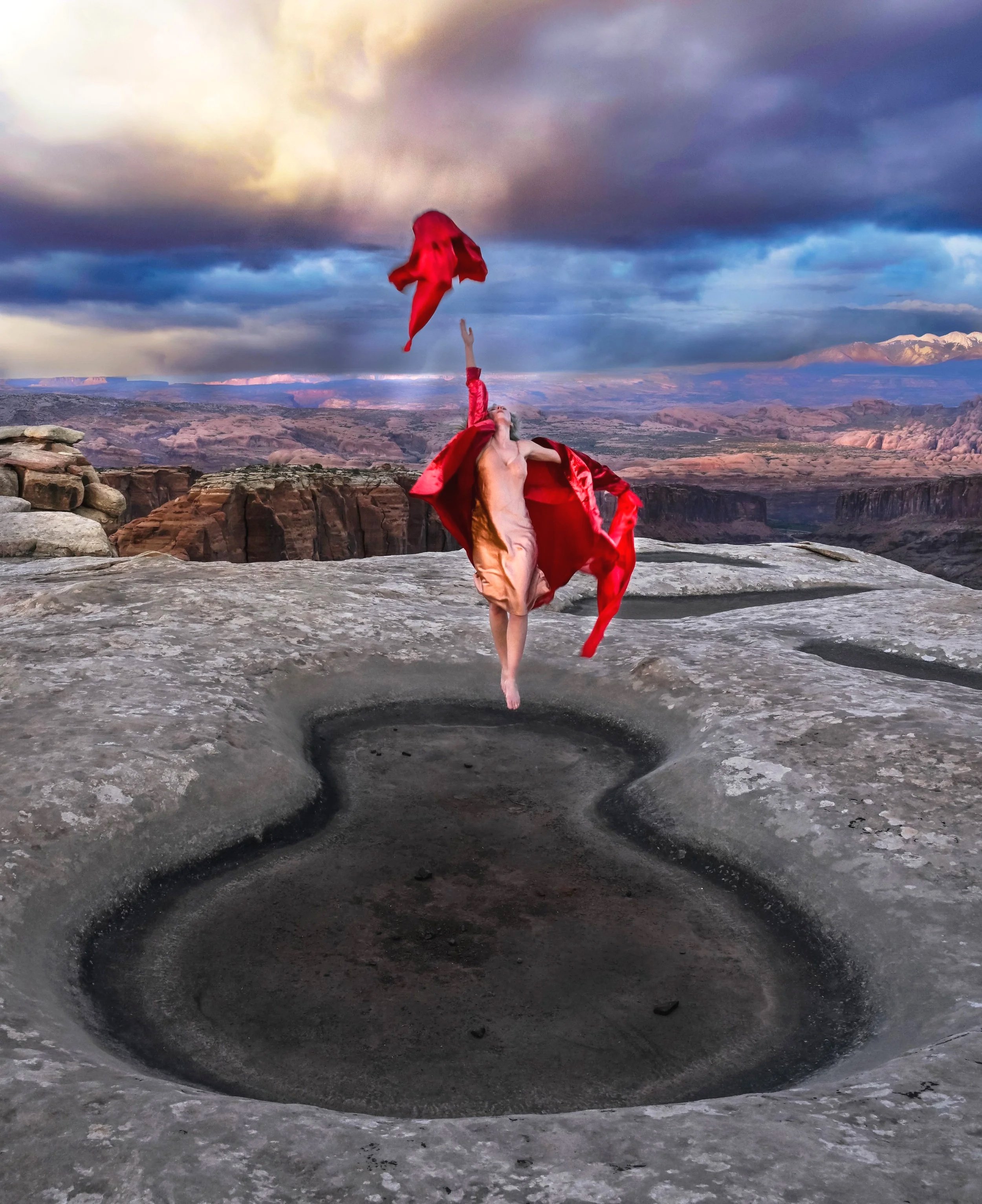 A woman in a beige dress and red jacket is jumping over a large circular hole in a rocky landscape with a dramatic sky and canyon scenery in the background.