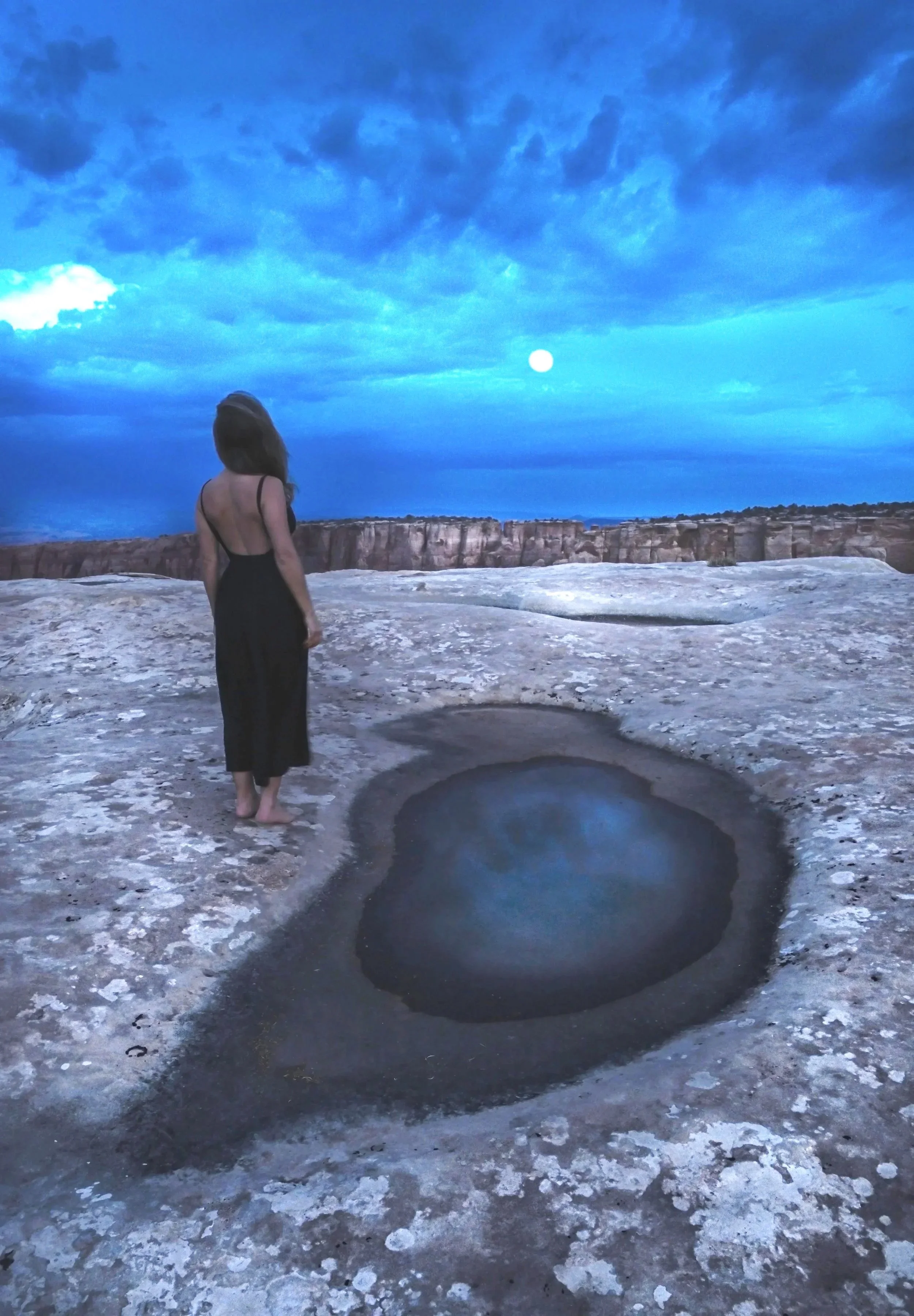 A woman in a black dress standing on a rocky landscape at dusk, looking at the moon in the sky. There is a small, round water hole in the ground nearby, reflecting the sky.