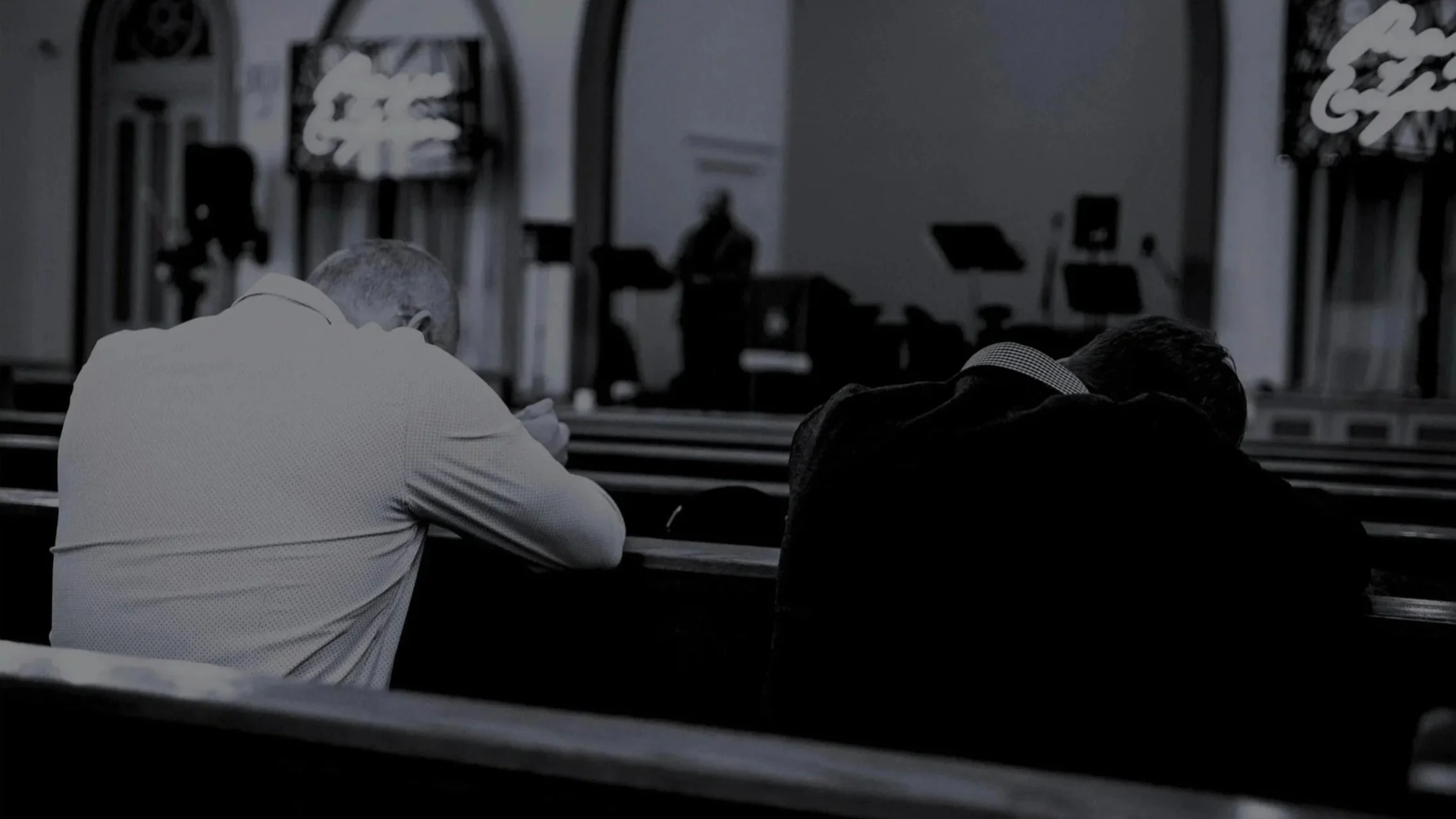 Two people sitting in church pews, leaning forward as if praying, with musical instruments and a speaker in the background.