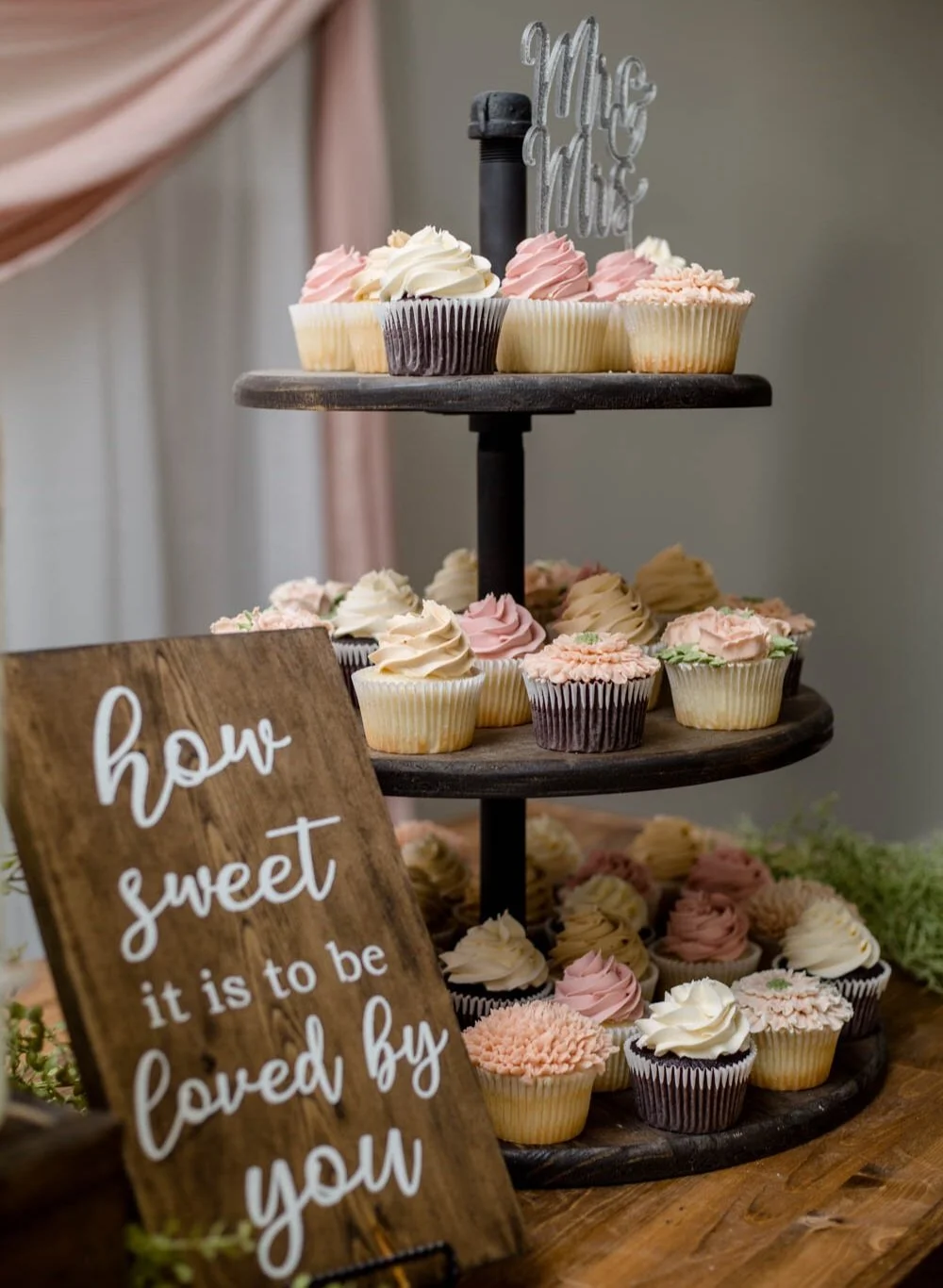A sign with the text "how sweet it is to be loved by you" sits in front of a wood dessert tower filled with different flavours of cupcakes