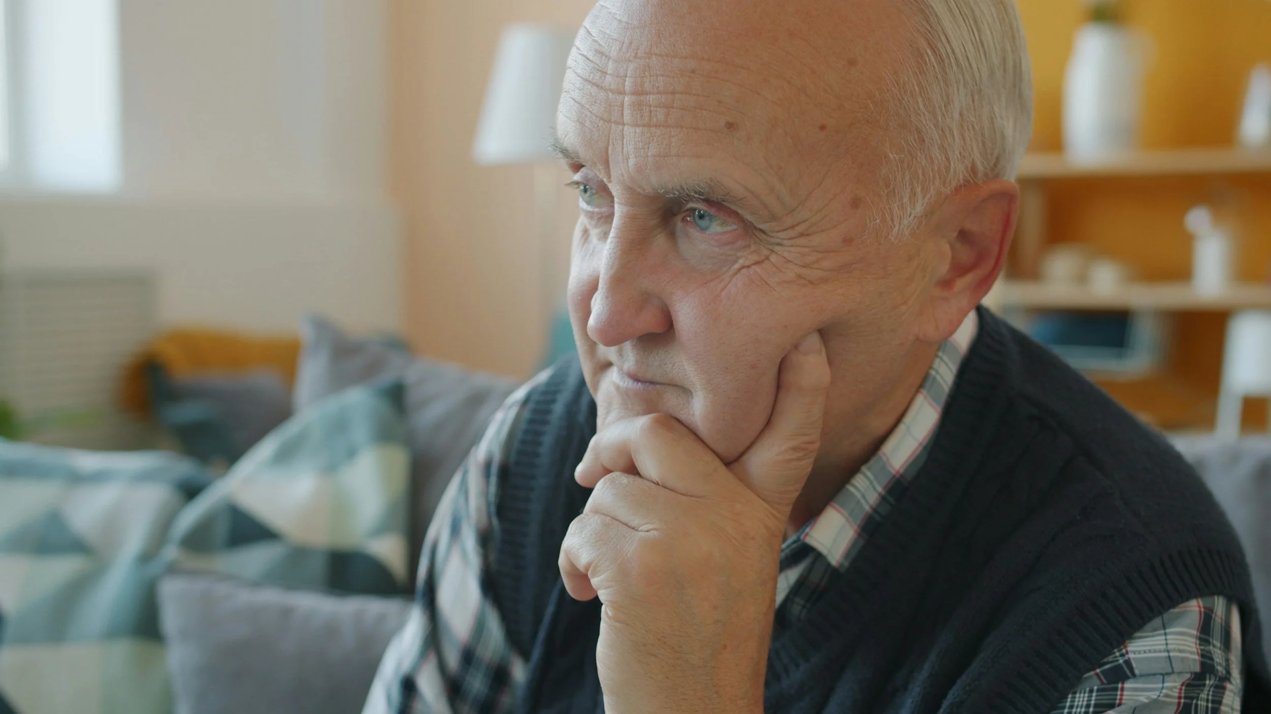 Close-up of an elderly man with light blue eyes, white hair, and a thoughtful expression, resting his chin on his hand indoors.