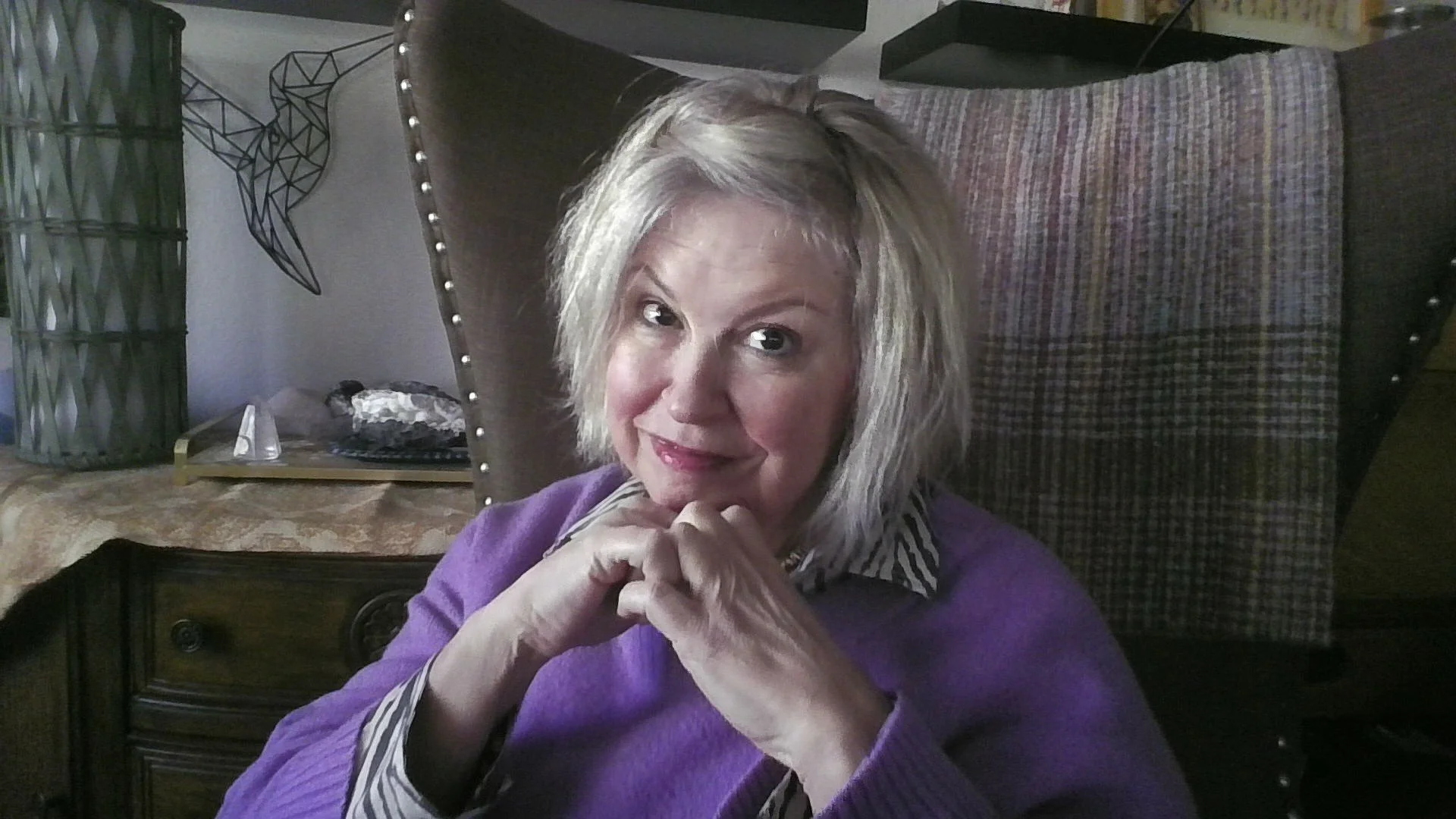 A woman with blonde hair in a layered style, sitting in a chair with studded and checkered fabric, wearing a purple sweater over a striped shirt, resting her chin on her clasped hands and smiling at the camera.