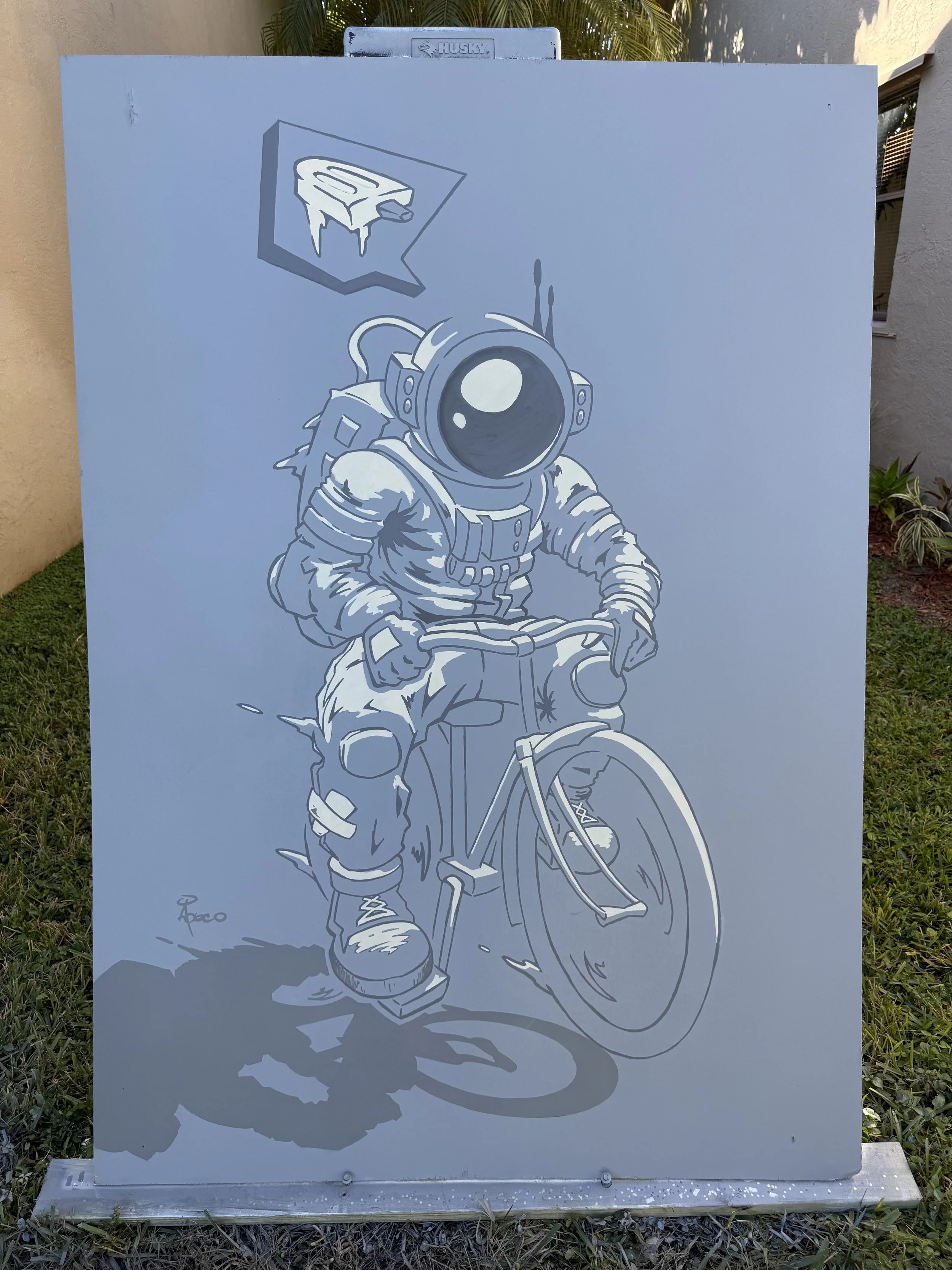 A monochrome outdoor mural of an astronaut riding a bicycle. The astronaut is wearing a spacesuit with a helmet, and the helmet's visor is reflective. There is a speech bubble above the astronaut's head containing an ice cream bar.