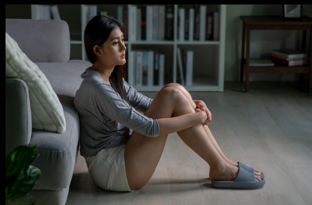 A woman sitting on the floor leaning against a couch, looking reflective and heavy-hearted
