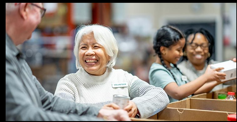 An older woman volunteer laughing warmly while working alongside others at a community food bank